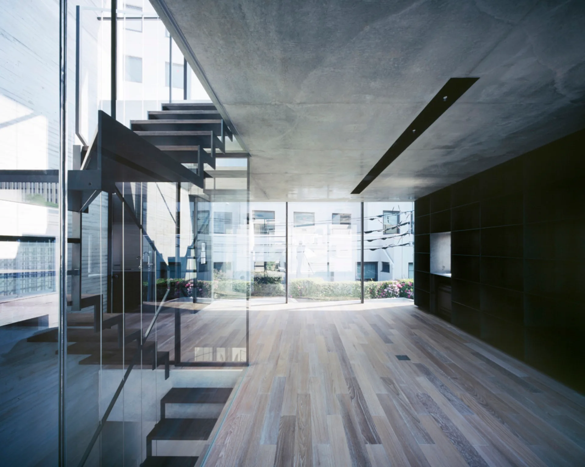 A modern minimalist interior features an open-riser wood staircase with a glass railing, light wood flooring, and a raw concrete ceiling.