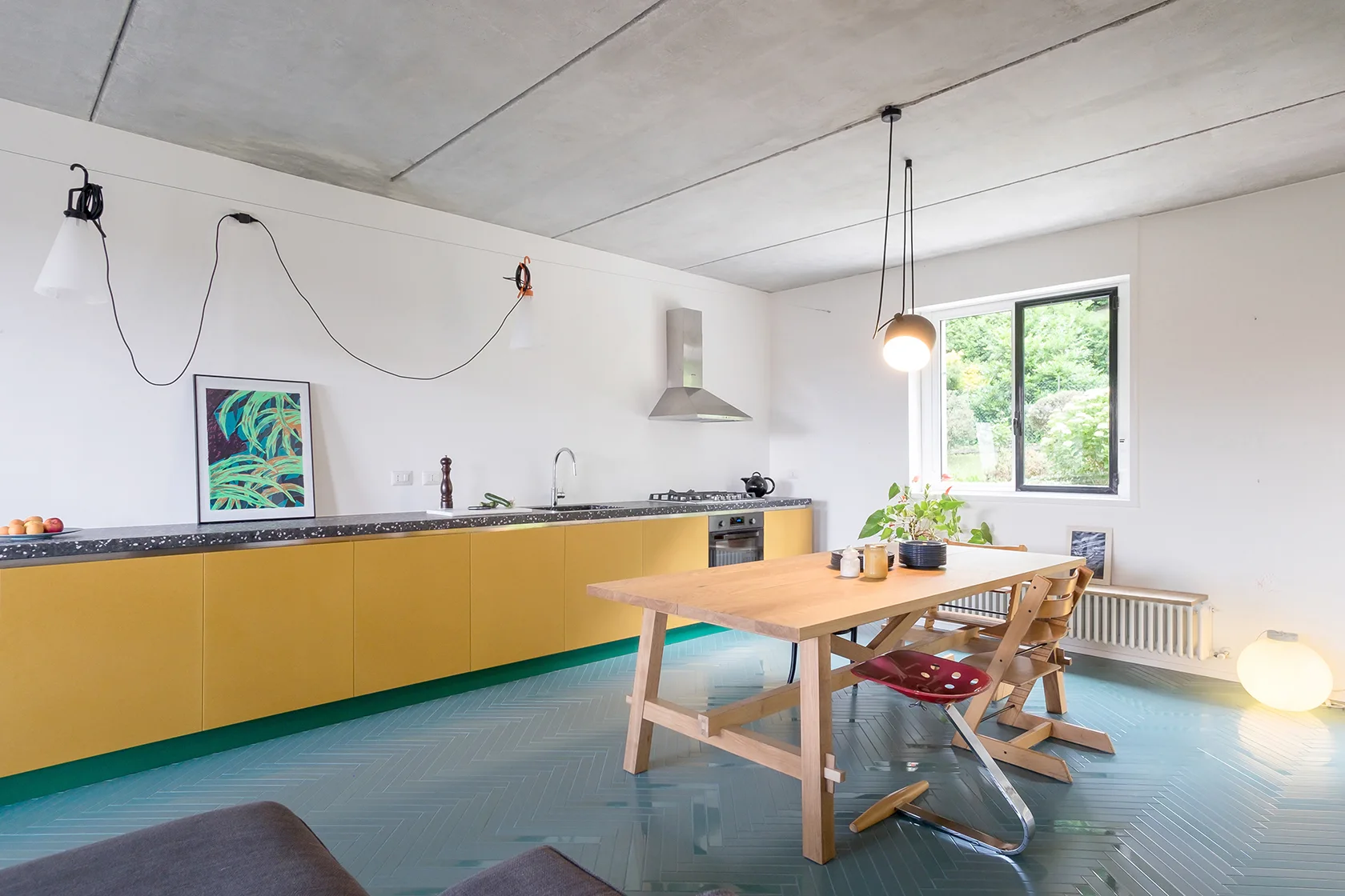 A modern kitchen features yellow base cabinets, a light wood dining table, blue-grey herringbone tile flooring, and an exposed concrete ceiling.