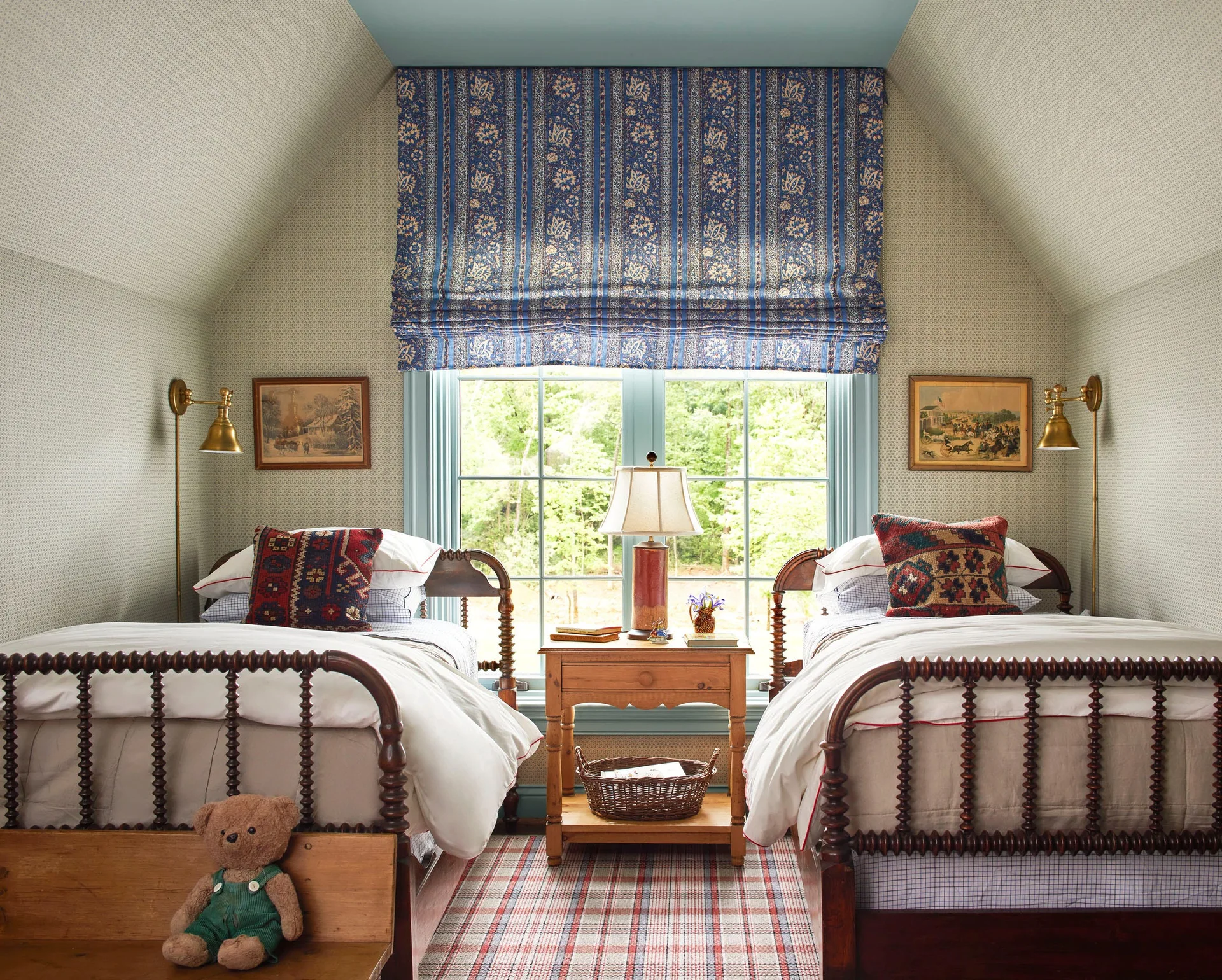A traditional cottage bedroom features two dark wooden spool beds flanking a central window decorated with a blue patterned roman shade.