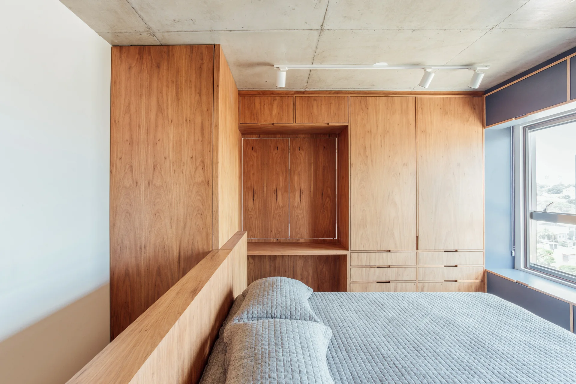 A modern bedroom with integrated wood cabinetry, a bed with gray quilted bedding, and an exposed concrete ceiling with track lighting.