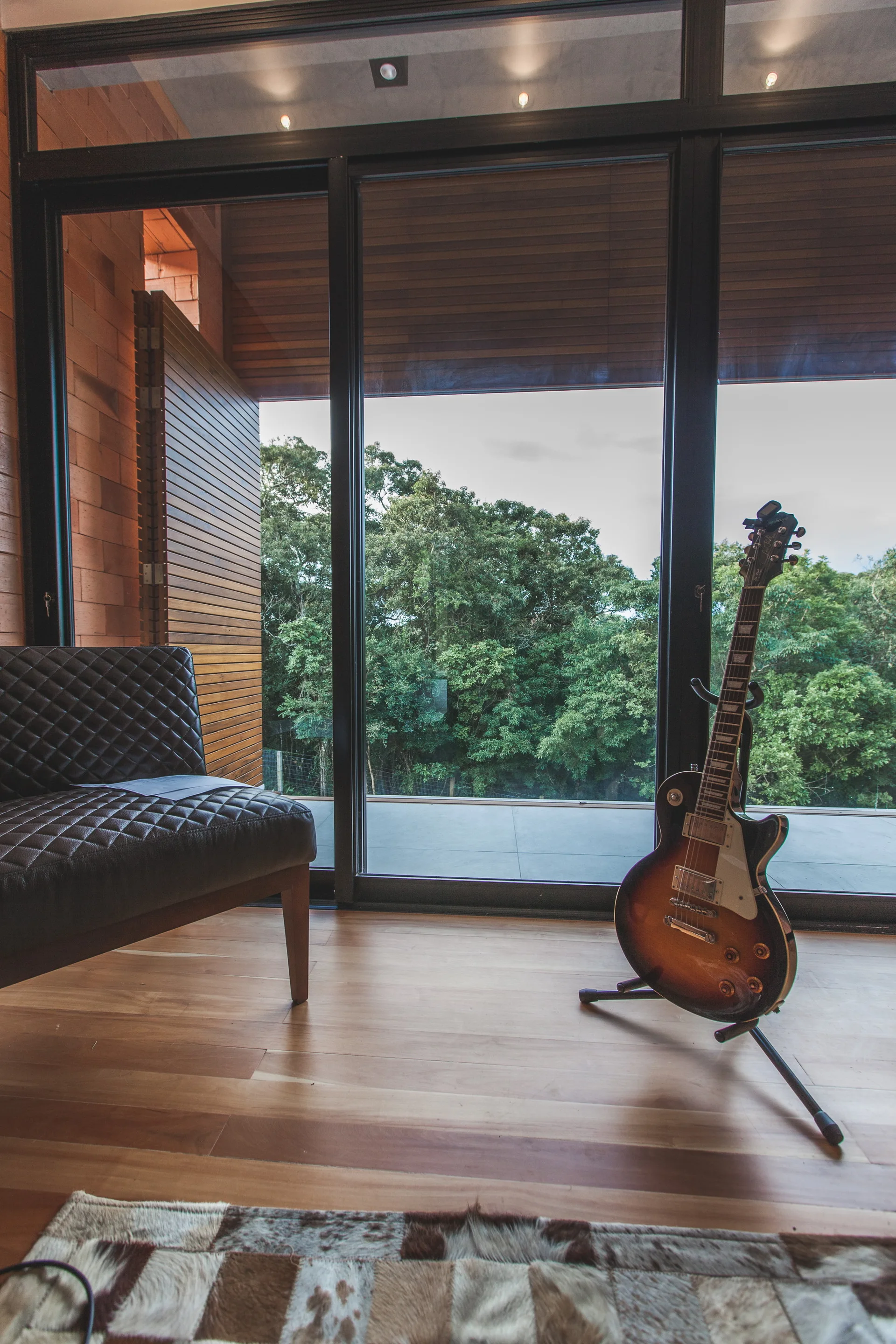 A quilted dark leather accent chair and an electric guitar on a stand sit on wood floors next to large glass sliding doors opening to a forest view.