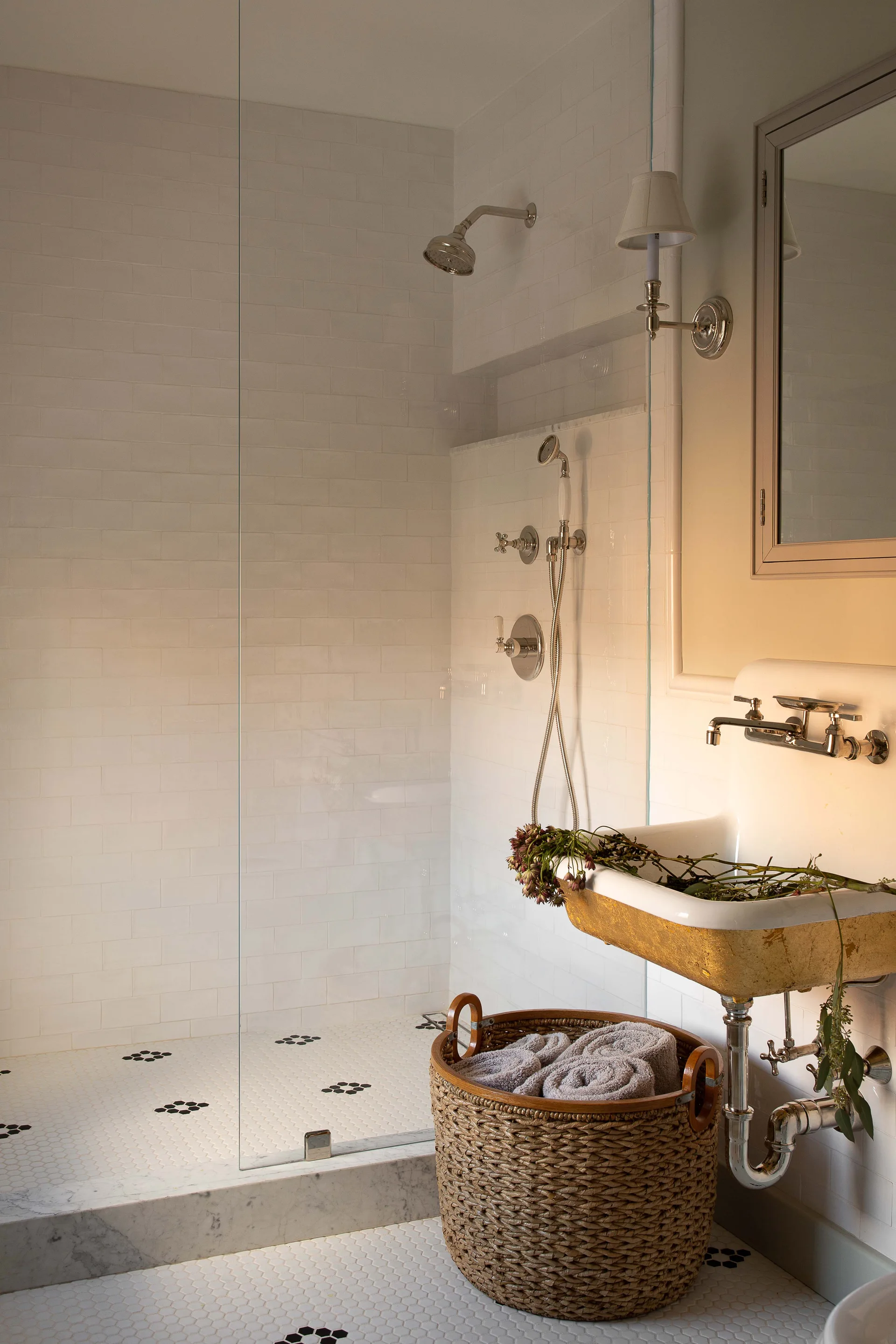 A white bathroom featuring a glass walk-in shower with subway tiles, a wall-mounted sink with a gold underside, and a wicker basket on a hexagonal tile floor.