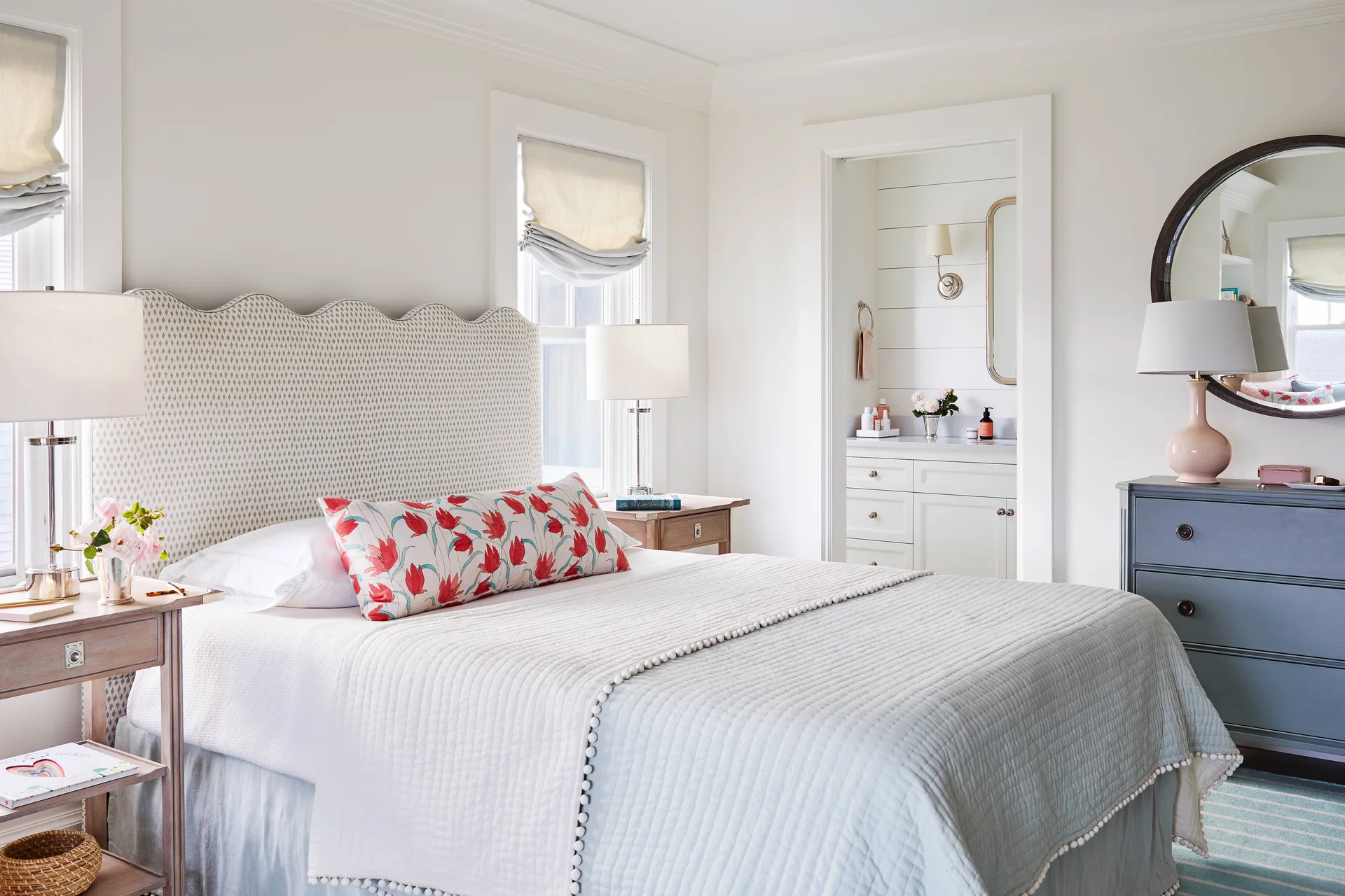 A bright bedroom features a patterned scalloped headboard, quilted bedding with pom-pom trim, and a doorway leading to a white shiplap bathroom.