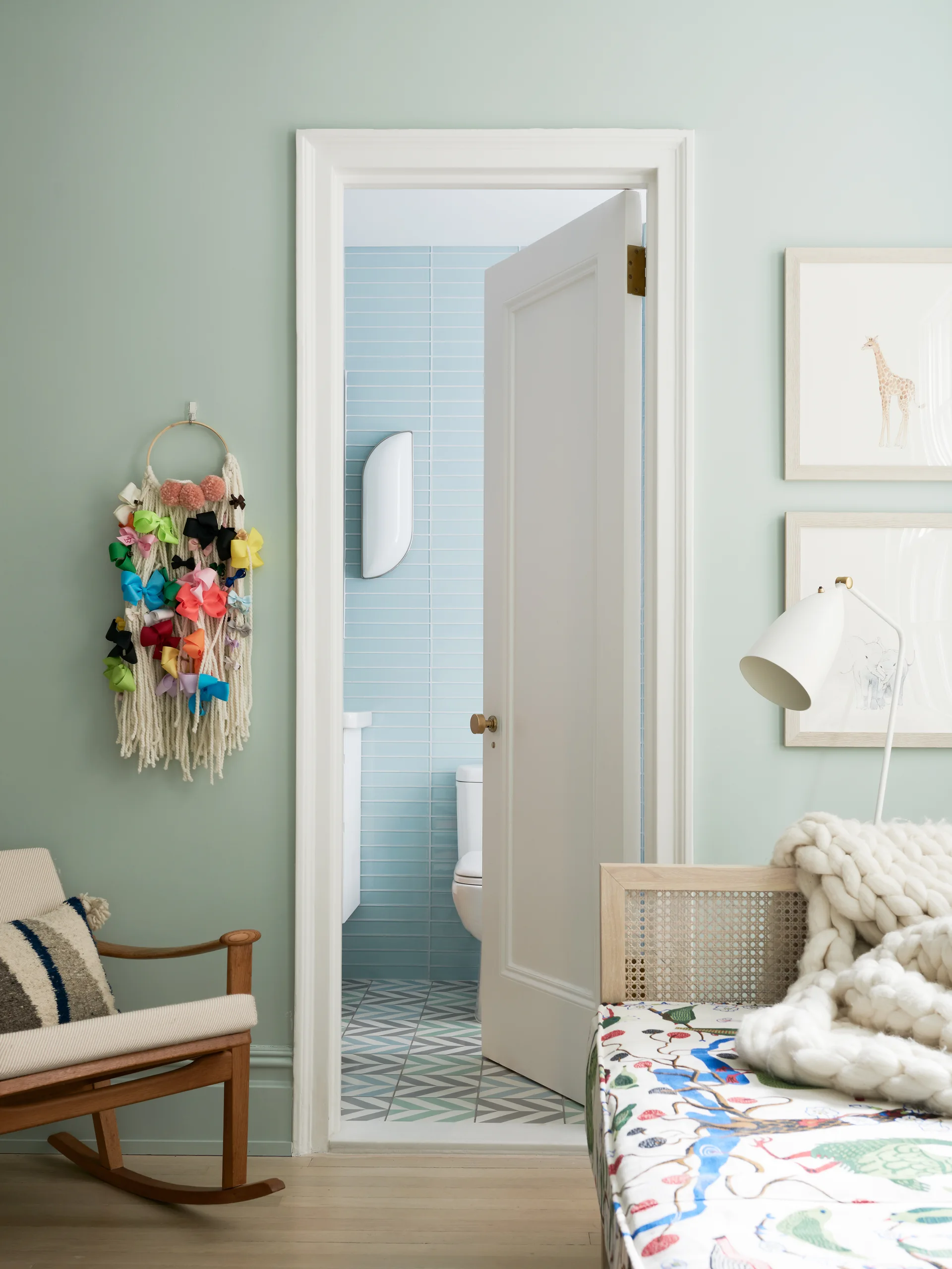 An open white door connects a sage green bedroom with a cane-back daybed to a bathroom featuring light blue wall tiles and geometric floor tiles.