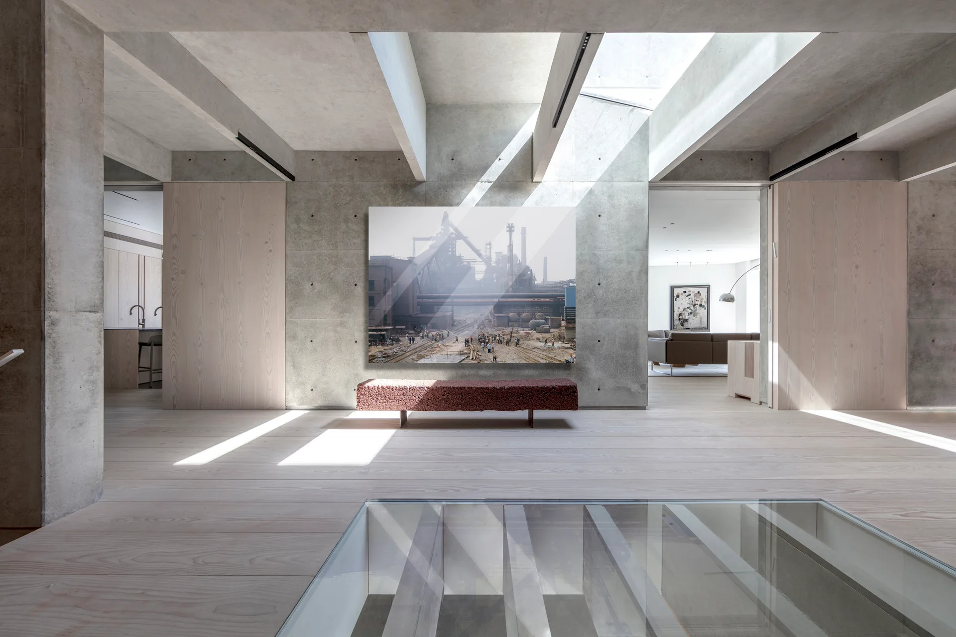 A minimalist hallway features exposed concrete walls, light wood flooring, a textured red bench under a large artwork, and light streaming from a skylight.