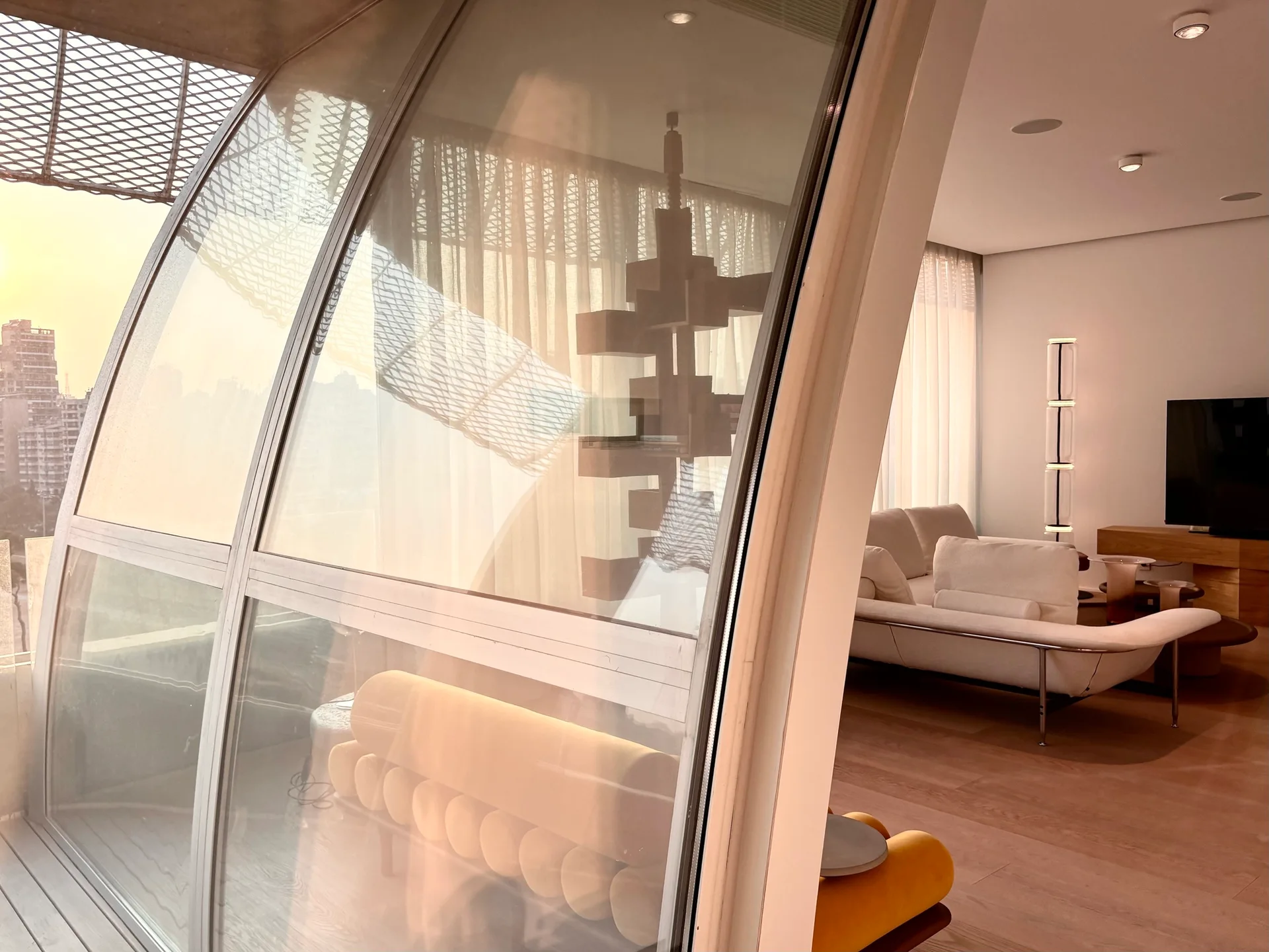 A modern living room with a white sofa and wood floors, seen through a large curved glass wall next to a yellow tufted bench.