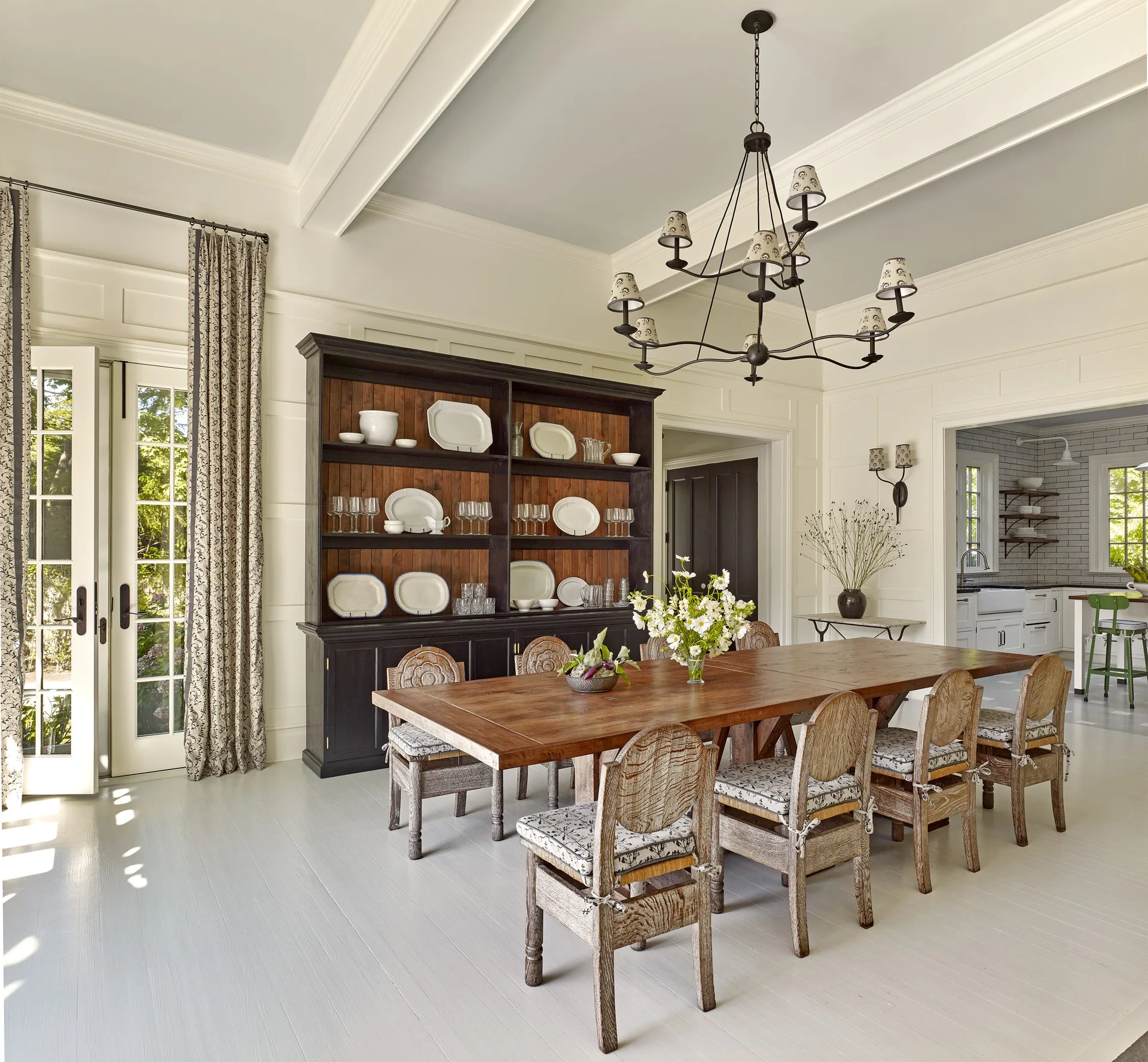 A wooden dining table and chairs sit on light grey painted floors in a room with a large black hutch displaying white dinnerware.