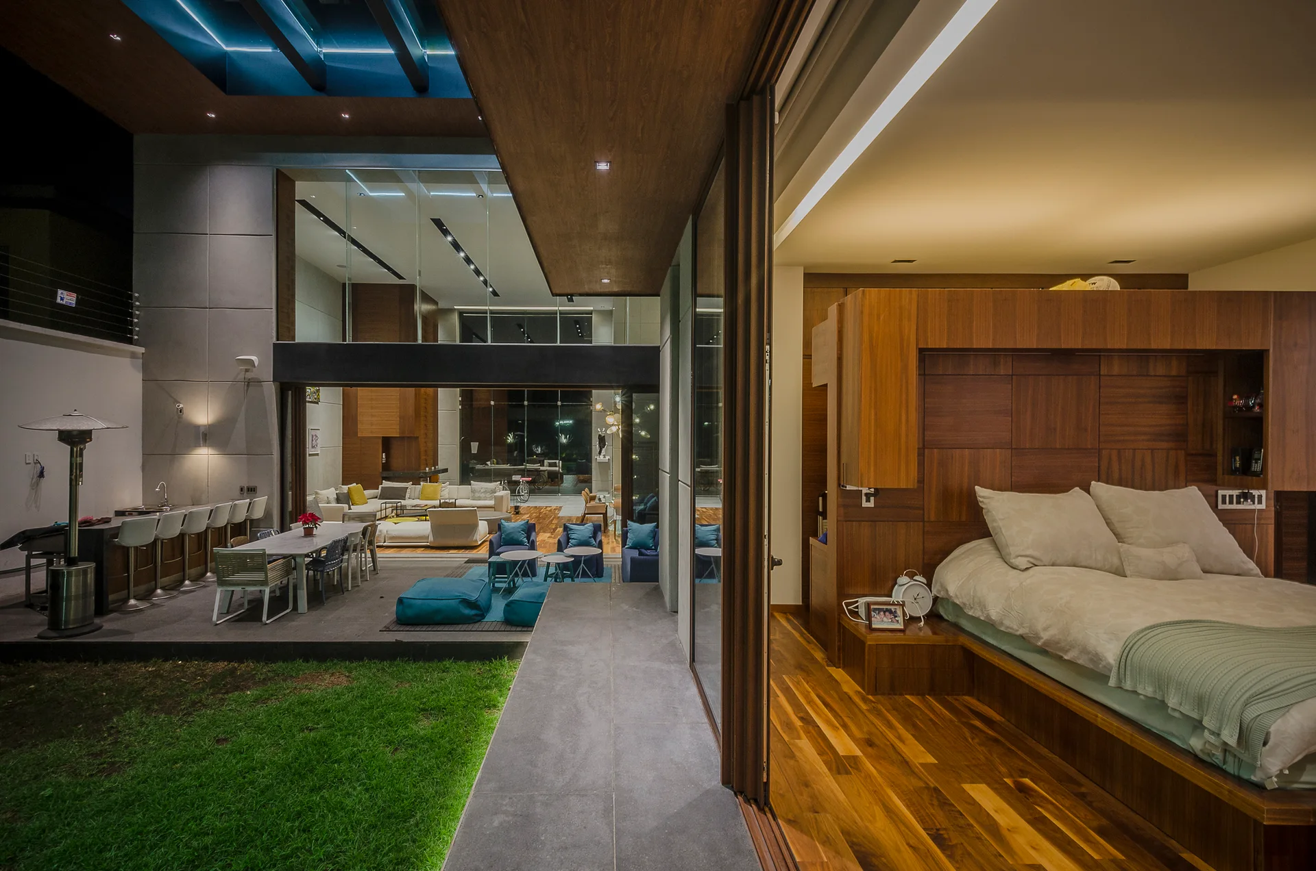 A modern bedroom with a custom wood platform bed and sliding glass doors that open to a furnished outdoor patio and lawn at night.
