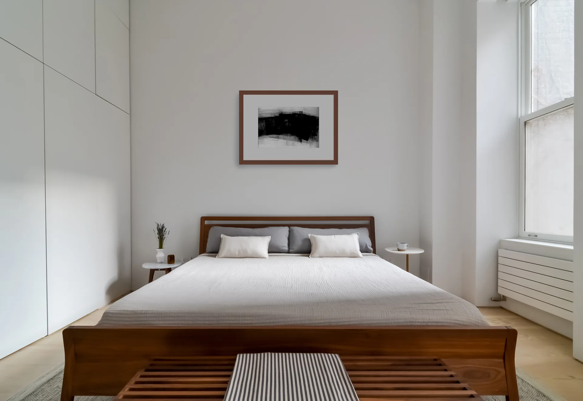 A minimalist bedroom featuring a wooden bed frame, seamless white built-in cabinets, and a framed abstract painting on the wall.
