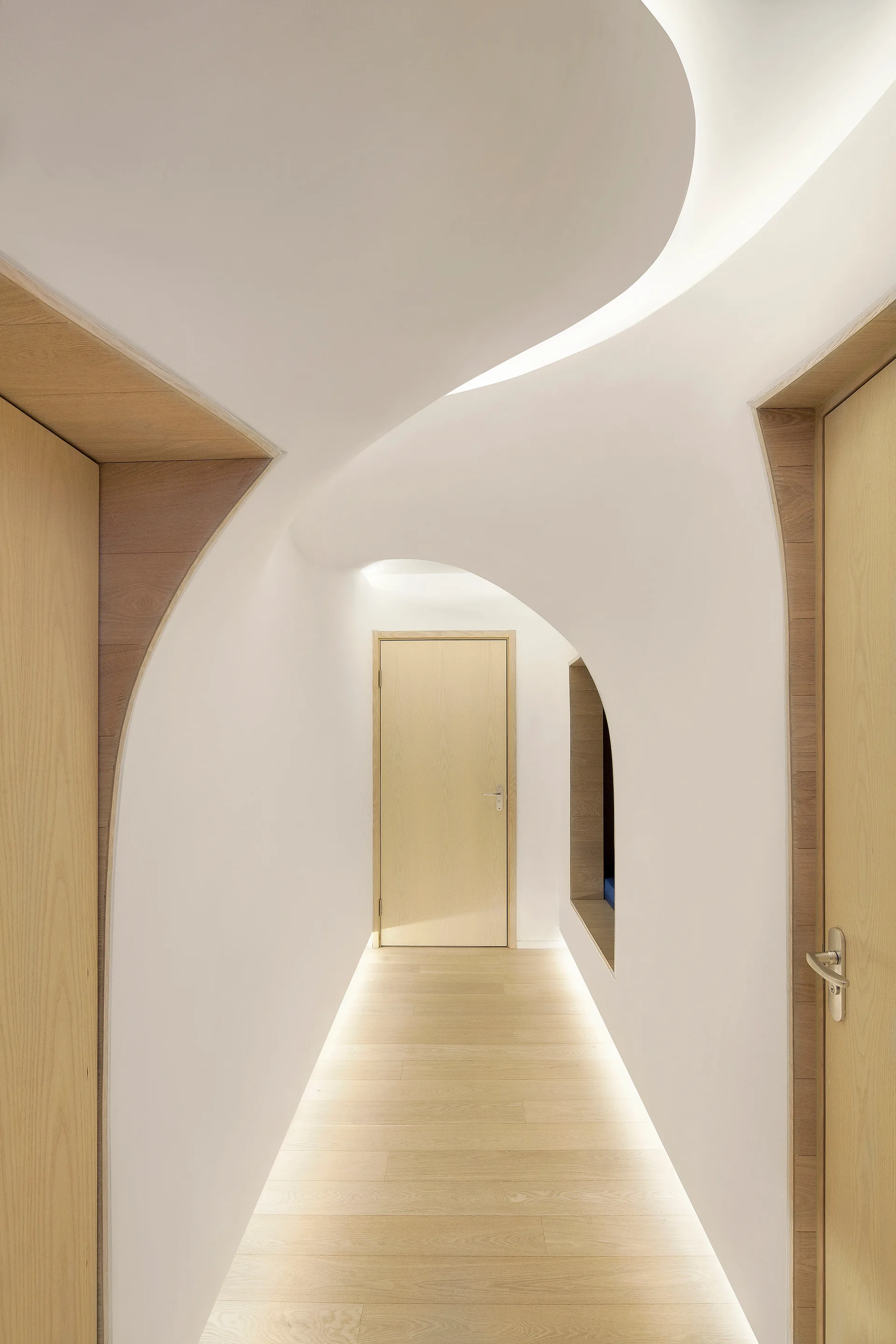 A minimalist hallway features sculpted, curved white walls, light wood doors, and integrated lighting glowing along the floor and ceiling.