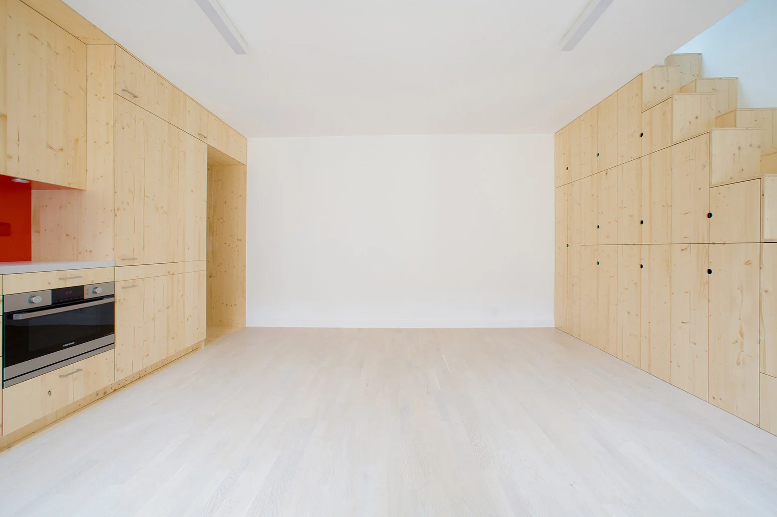 A minimalist room features light-toned wood cabinetry on both walls, with a built-in kitchen oven on the left and an integrated staircase on the right.