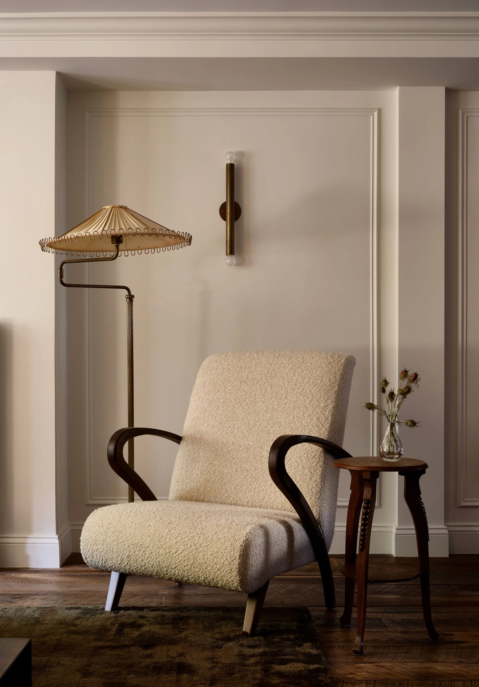 A cream textured armchair sits next to a small wooden side table and a brass floor lamp against a pale wall with decorative panel molding.