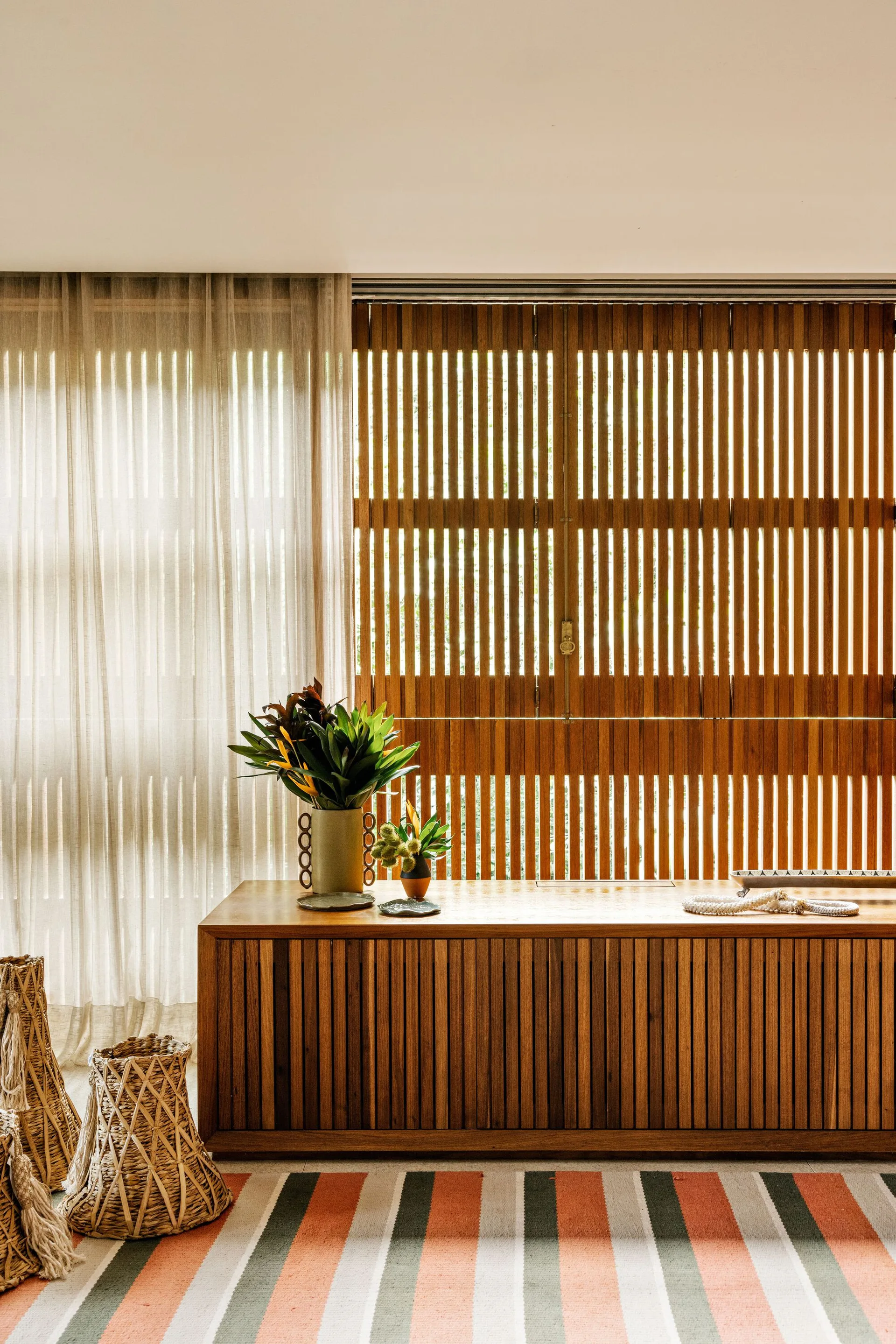 A low wooden slatted cabinet sits beneath a matching slatted window screen, flanked by a sheer linen curtain and woven floor baskets.