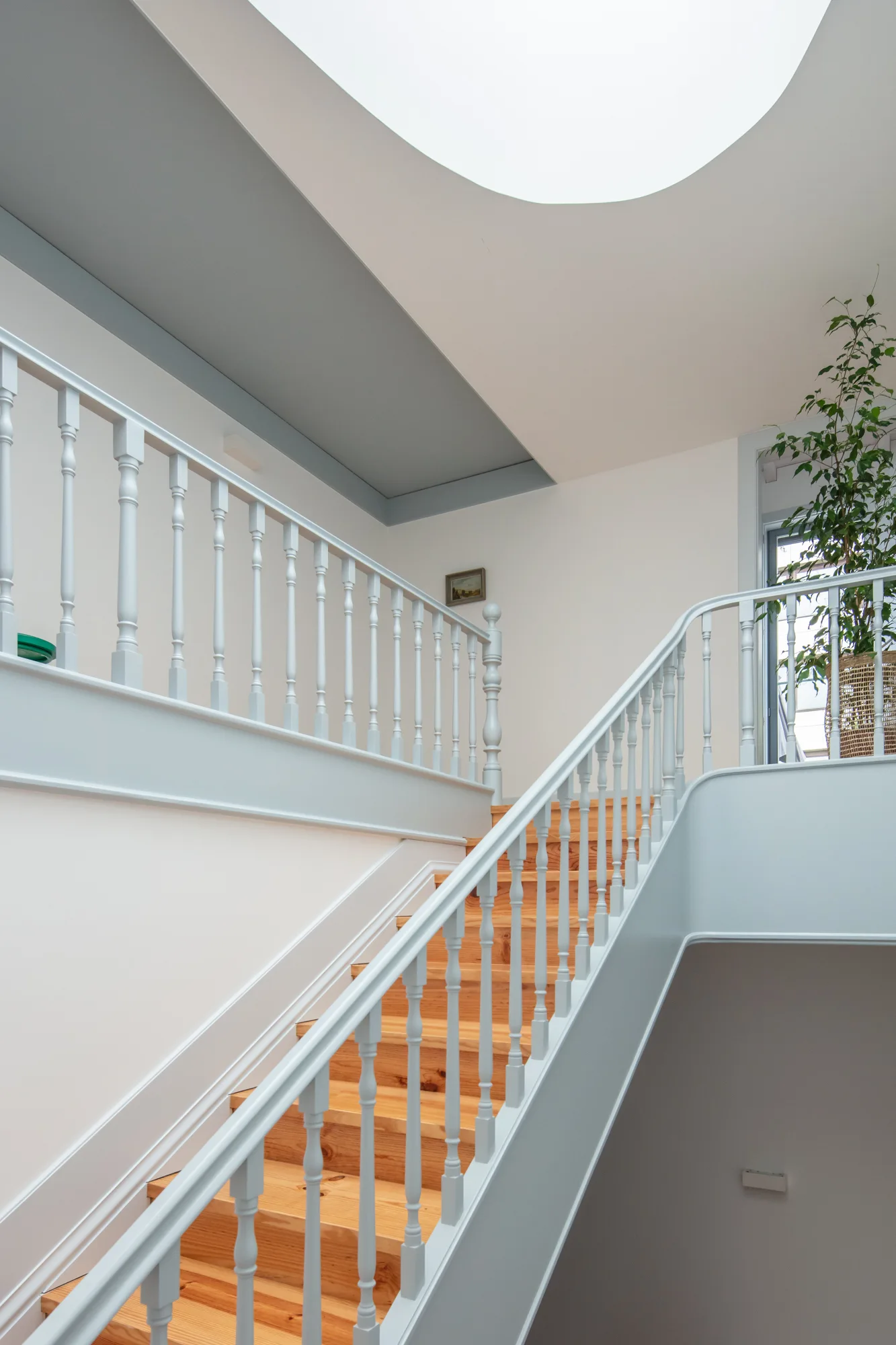 A wooden staircase with natural treads and pale blue painted railings rises toward a circular skylight in a white hallway.
