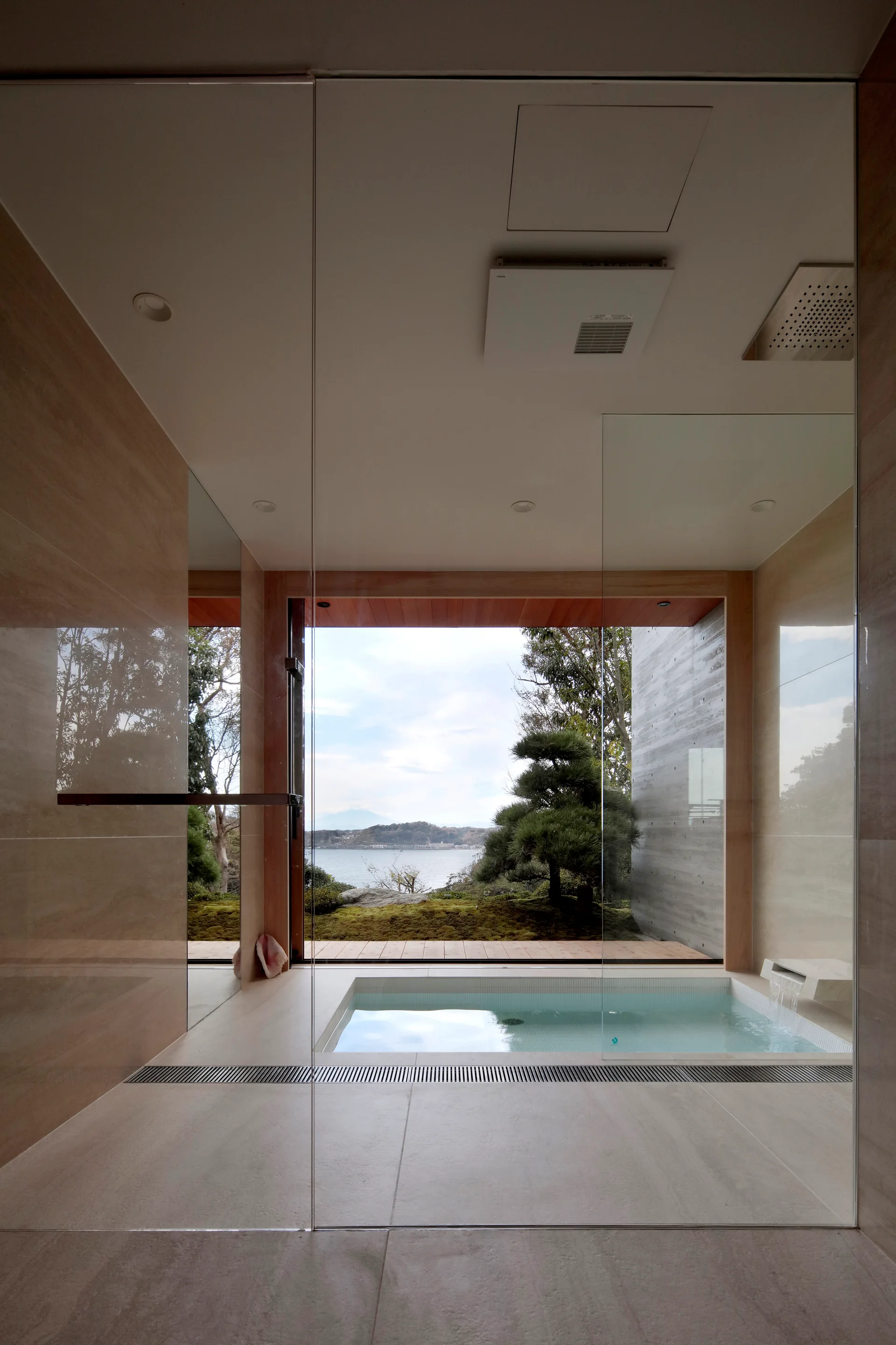A minimalist spa-like bathroom features a sunken bathtub and frameless glass partition walls next to a large window overlooking trees and water.