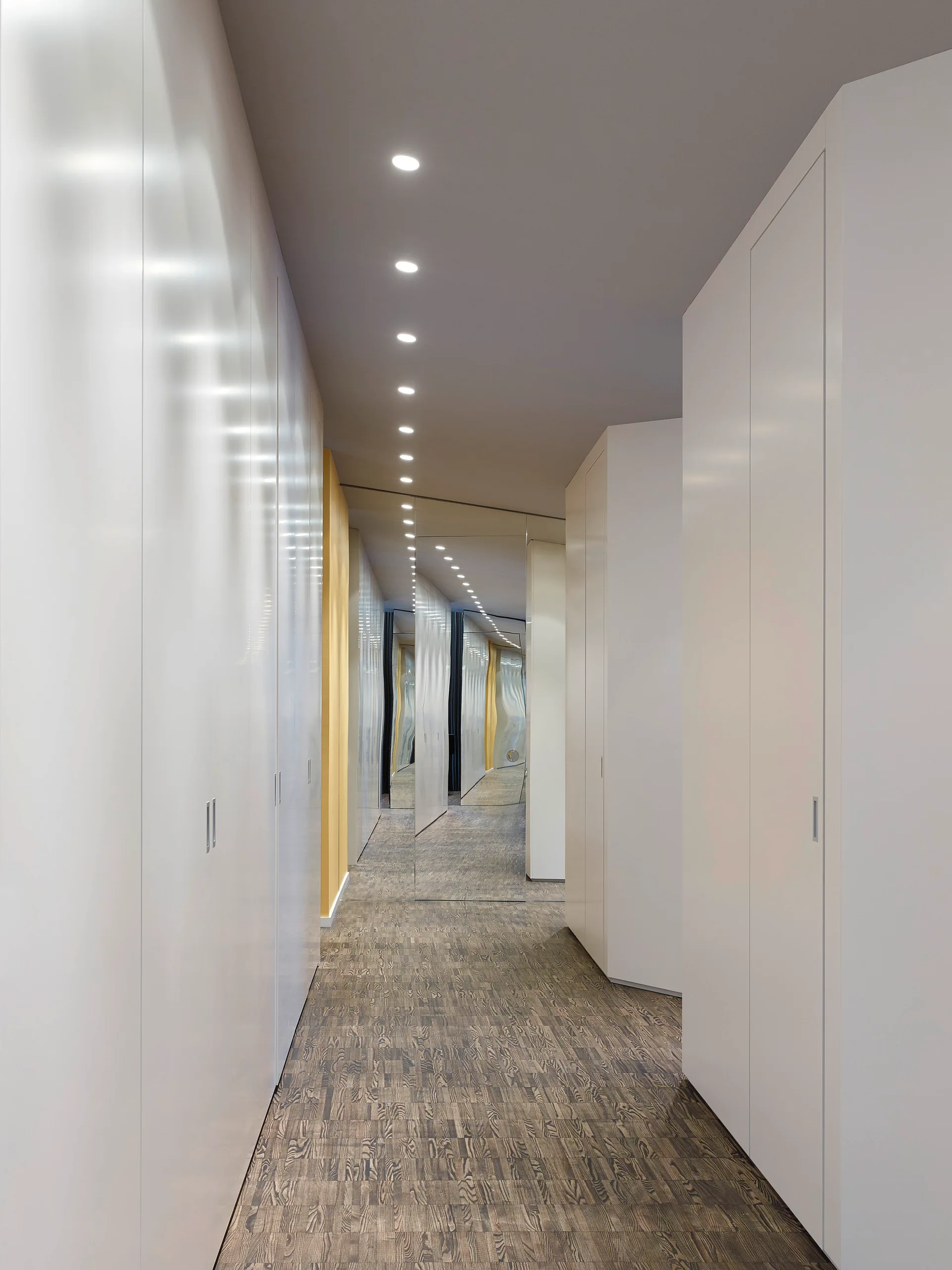 A modern hallway features glossy white built-in cabinets on both sides, a mirrored end wall, and recessed ceiling lights over textured wood flooring.