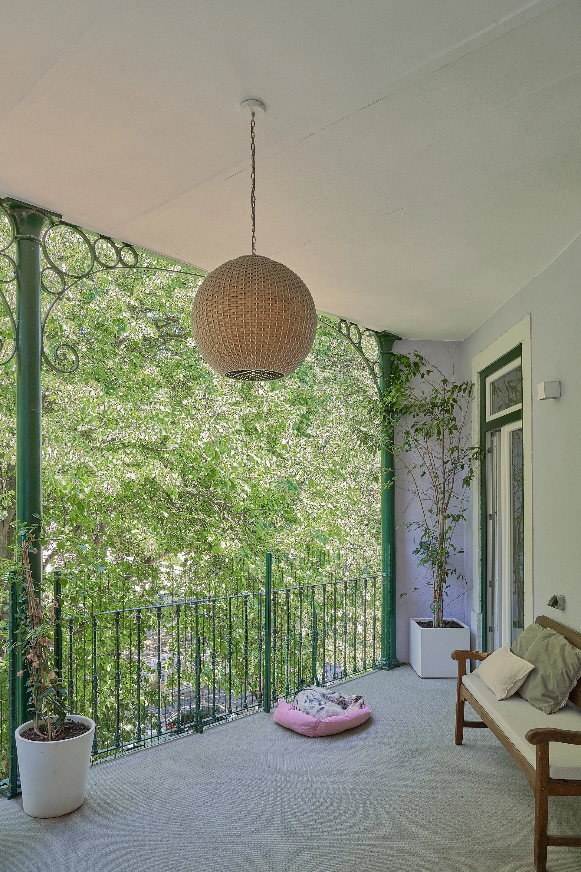 A covered balcony features green ornate iron railings and columns, a large spherical woven pendant light, and a wooden bench.