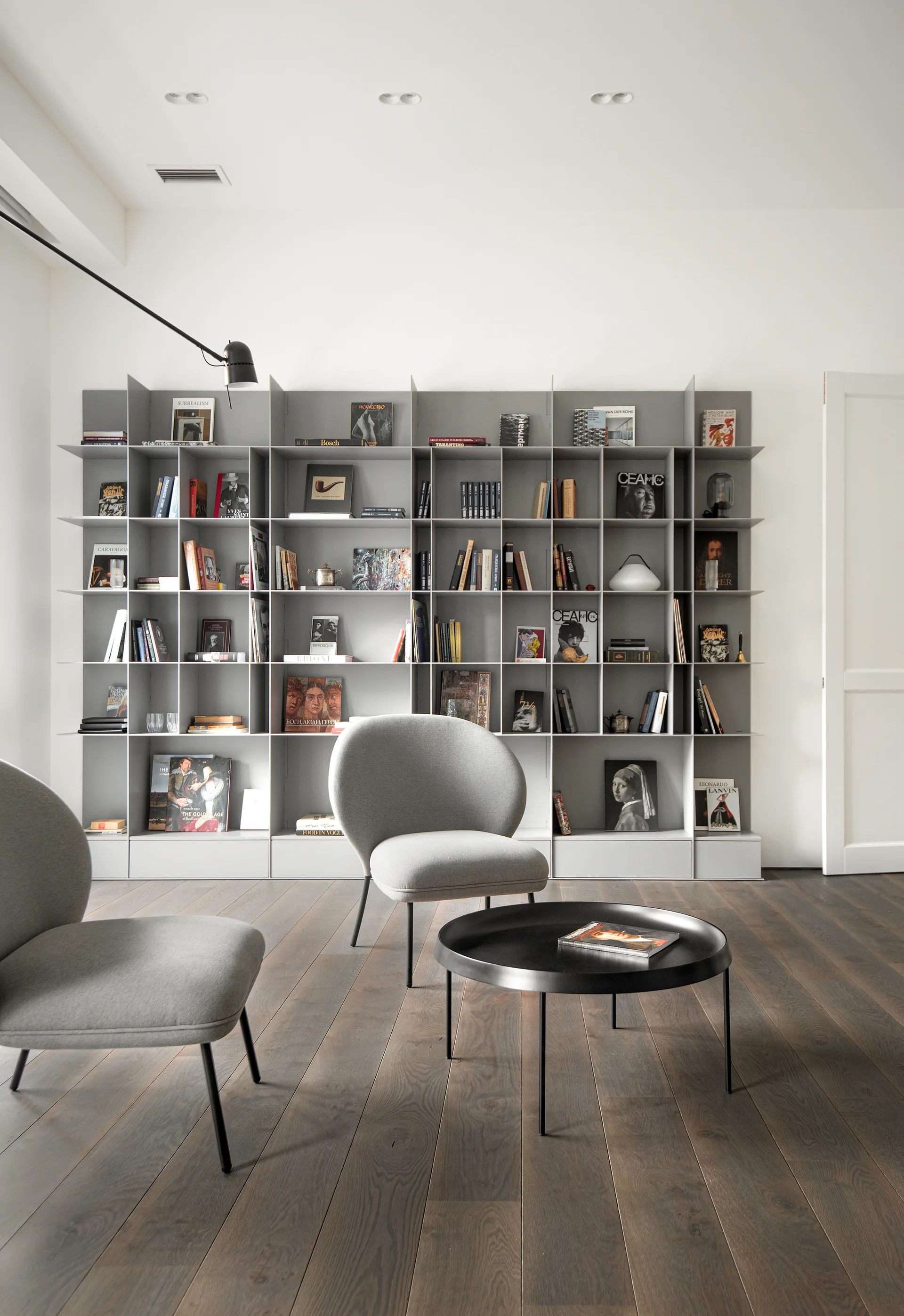 Two grey upholstered lounge chairs and a round black coffee table stand in front of a tall grey grid bookshelf on dark wood flooring.