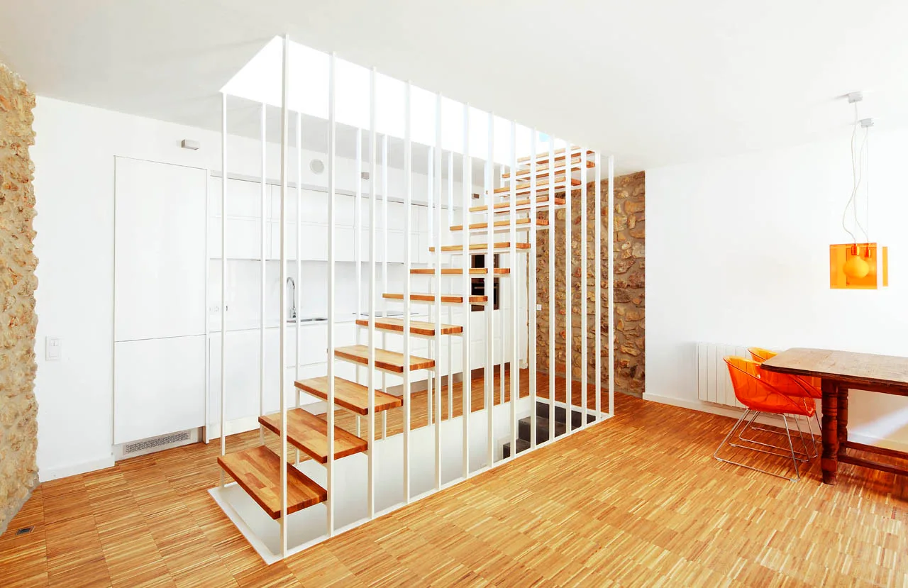 A modern open-plan room features a floating wooden staircase with floor-to-ceiling white metal railings, set against a natural stone accent wall.