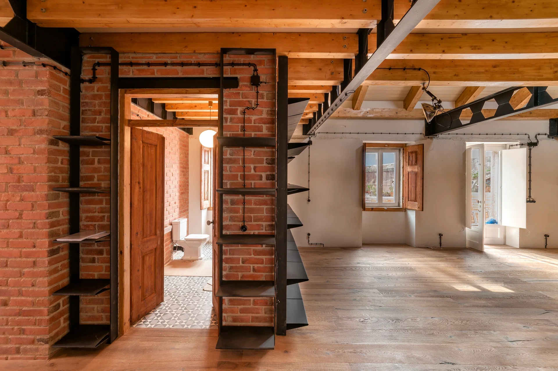 An industrial loft interior featuring exposed red brick walls, wood-beam ceilings, black metal shelves, and a doorway leading to a tiled bathroom.
