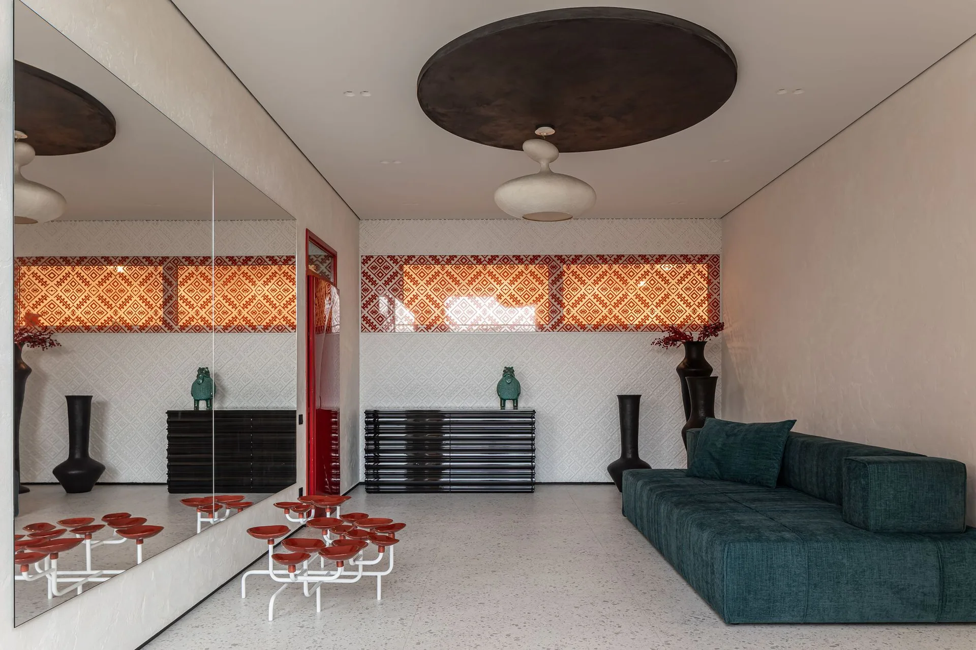 A contemporary living room with a dark green floor sofa, a large wall mirror, textured geometric white walls, and a cluster of red-topped stools.