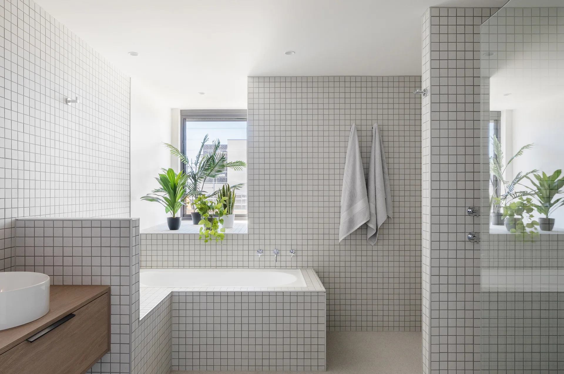 A minimalist bathroom features white grid-tiled walls and tub surround, a floating wood vanity with a vessel sink, and potted plants on a window ledge.