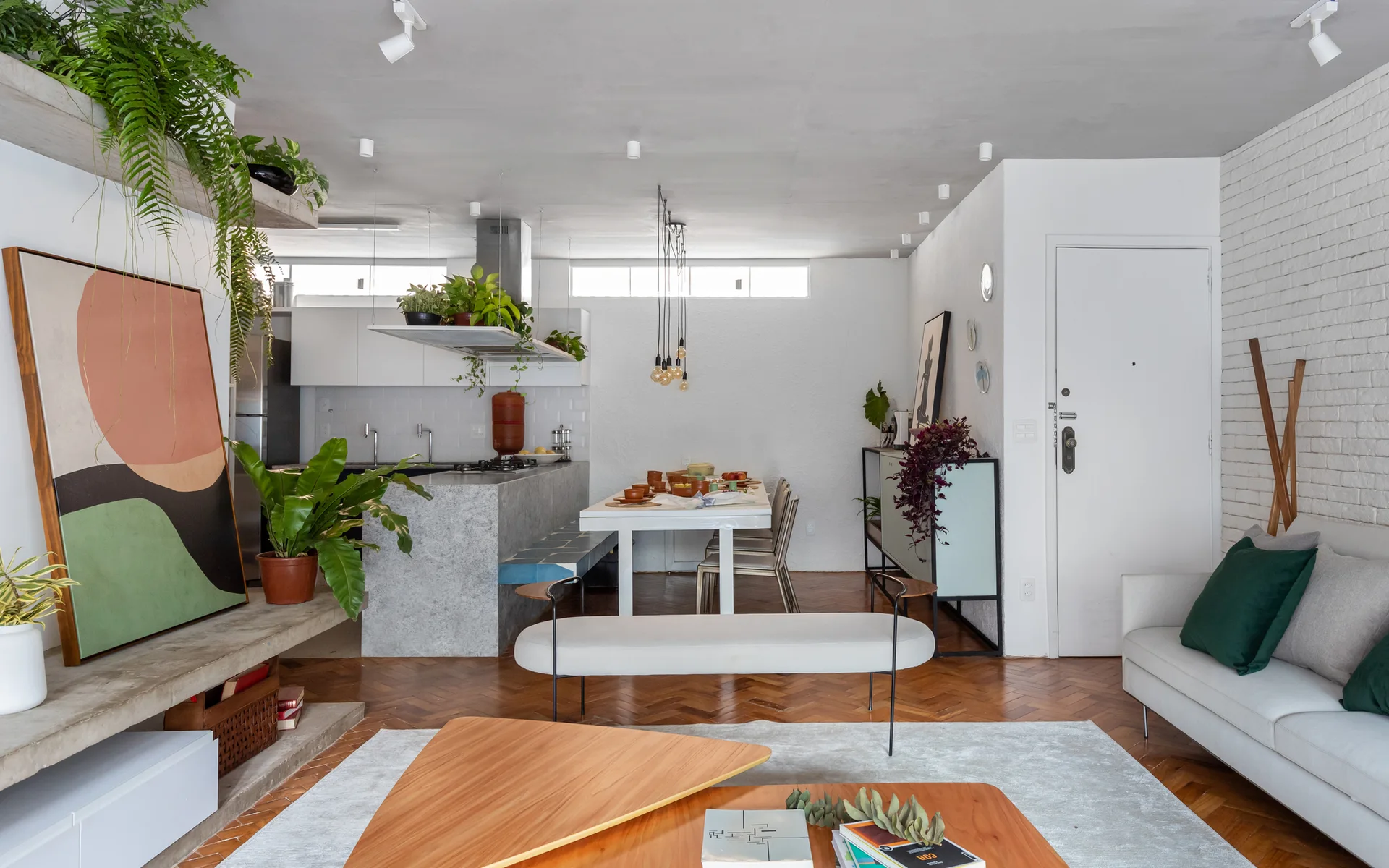 An open-plan living room and kitchen features herringbone wood floors, a white brick wall, a gray stone kitchen island, and green plants.