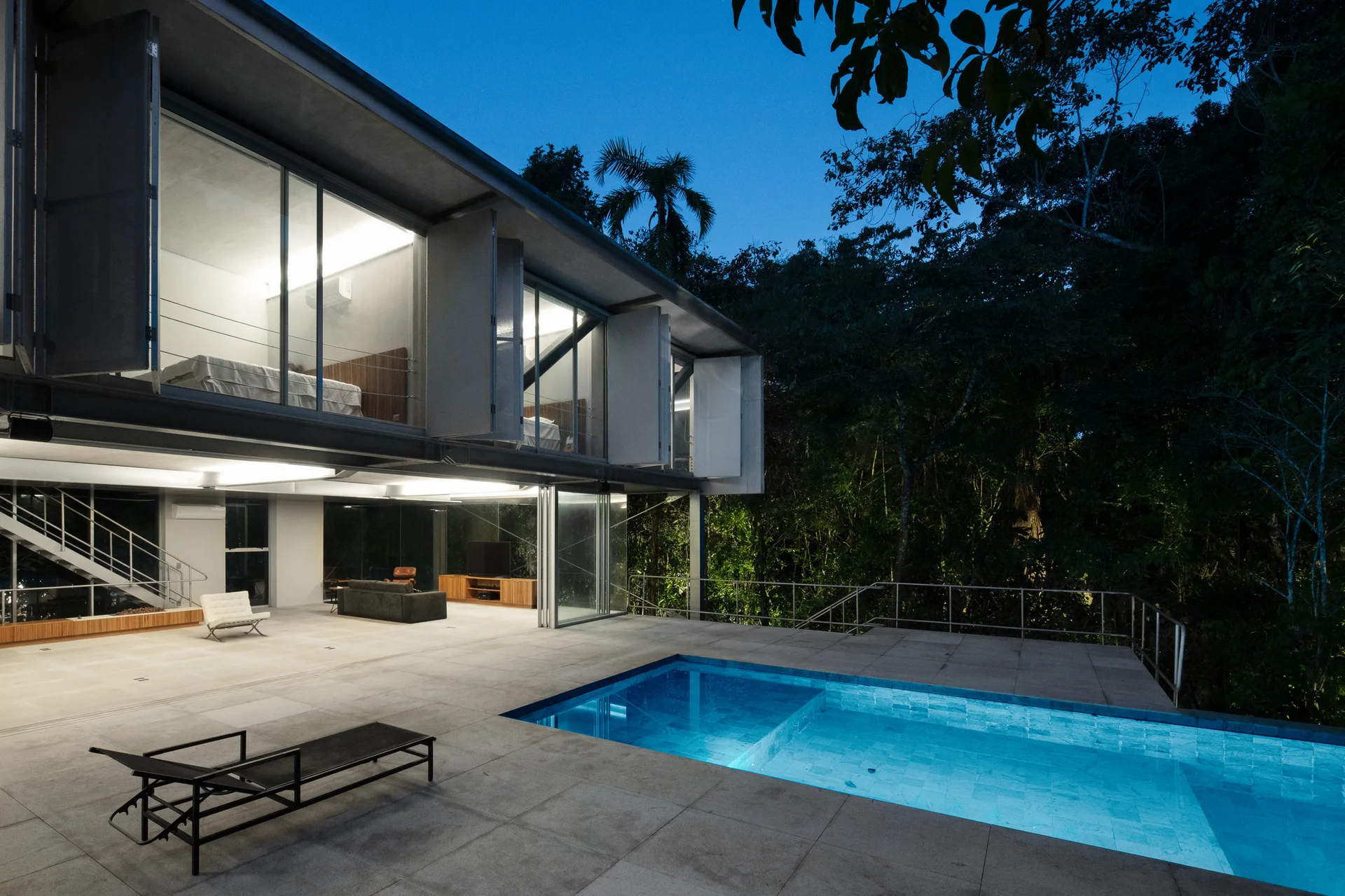 A modern two-story concrete residence with floor-to-ceiling glass walls overlooks a rectangular swimming pool on a grey stone terrace at dusk.