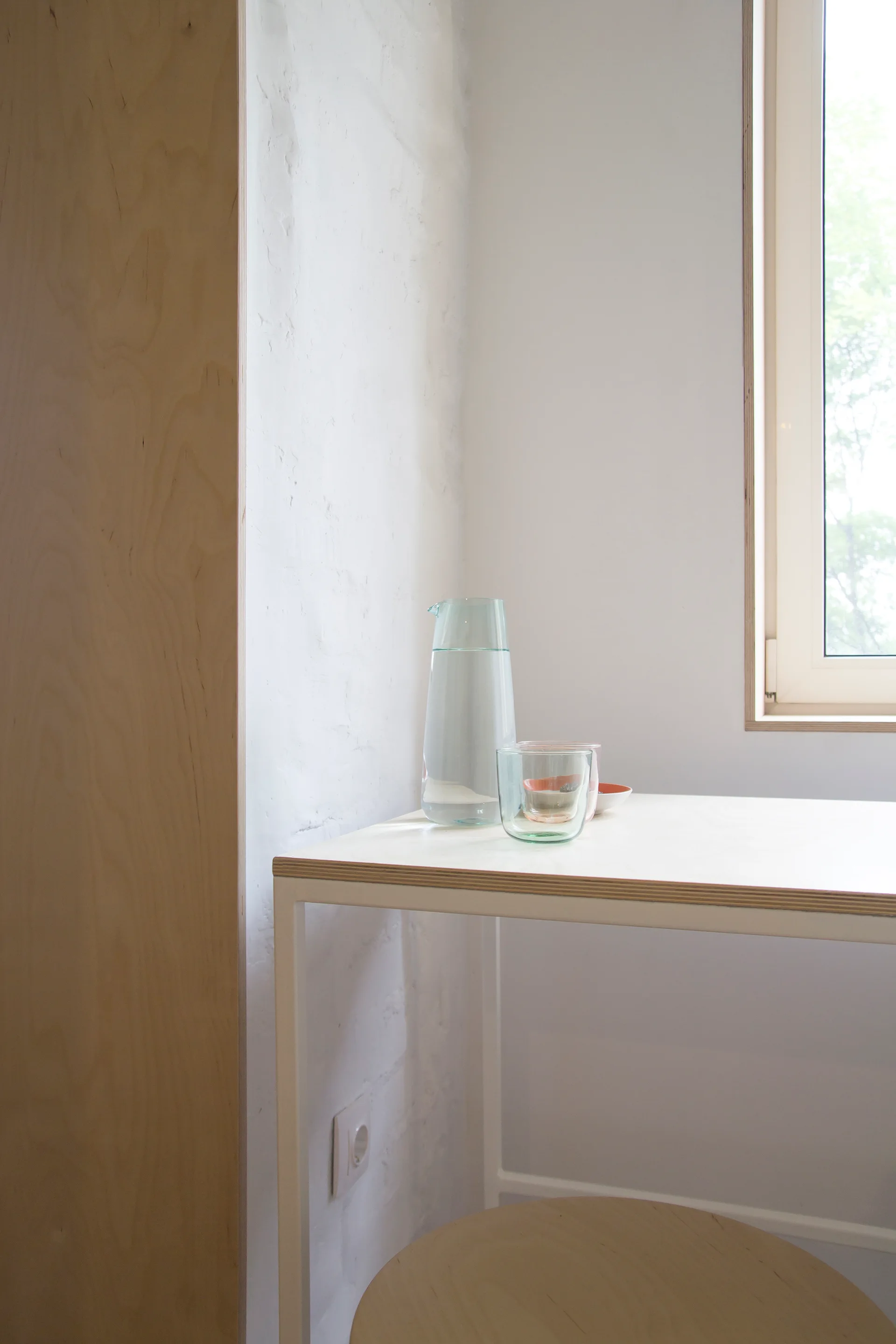 A white table with a glass water carafe sits next to a light wood window frame and a painted brick wall.