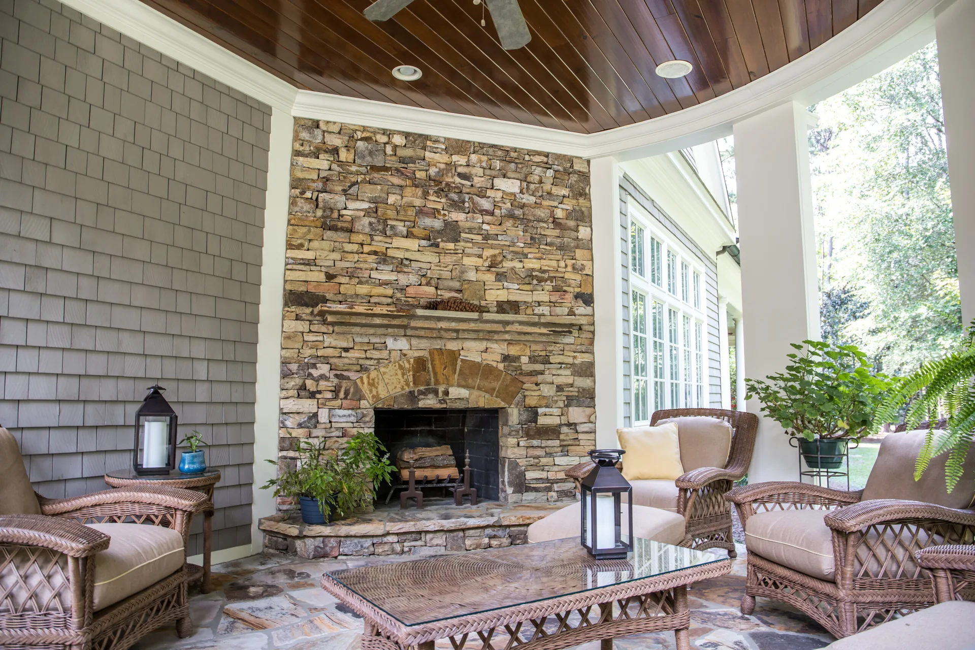 A covered patio with flagstone flooring features a stacked stone fireplace, wicker armchairs, a glass-top coffee table, and a polished wood ceiling.