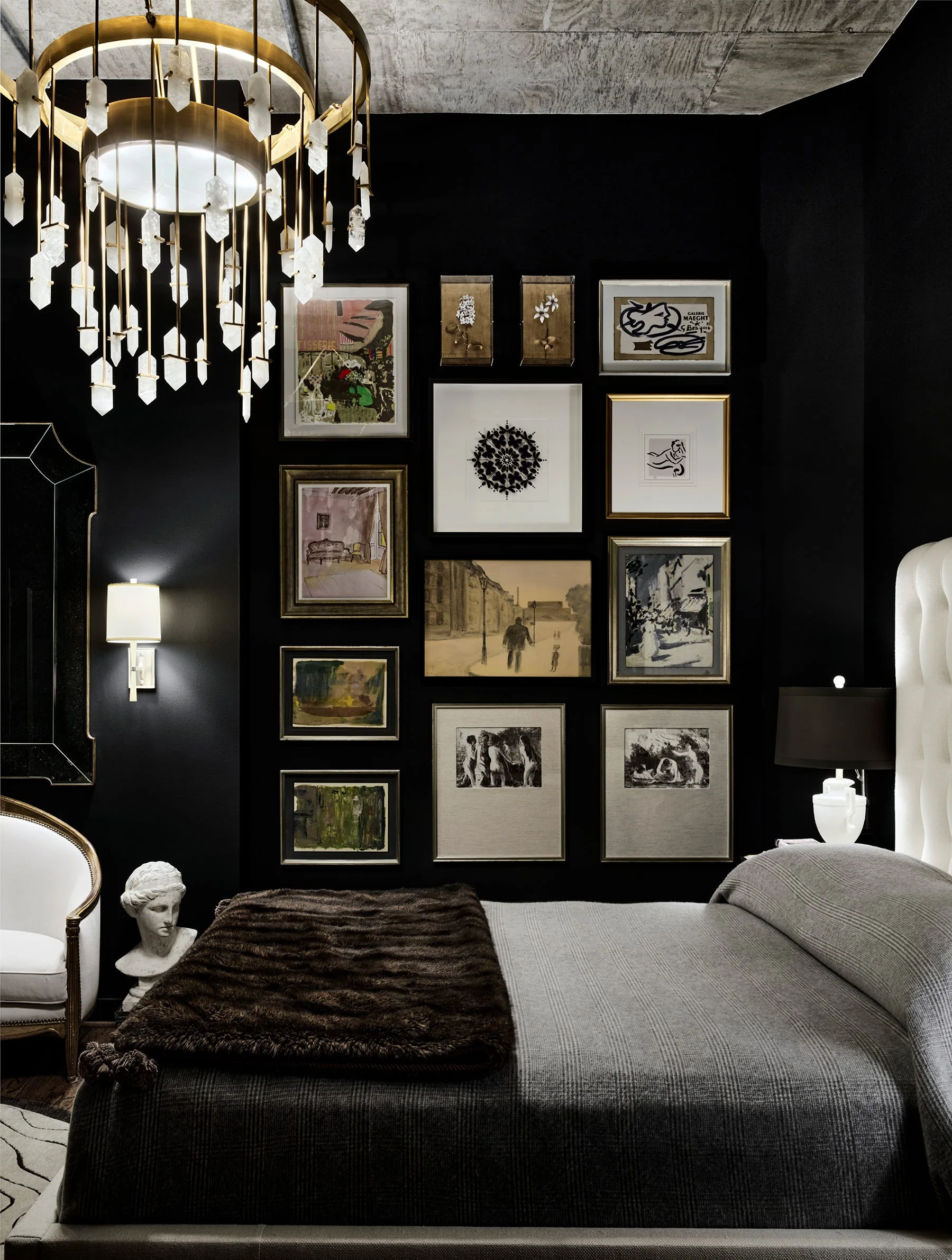A bedroom with dark black walls, a gallery art wall, a crystal chandelier, a bed with grey bedding, and a raw concrete ceiling.