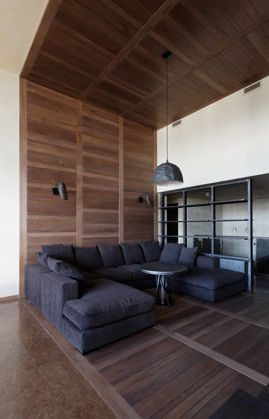 A dark charcoal sectional sofa sits under a wood-paneled ceiling and matching wall accent in a modern living room with a round metal coffee table.