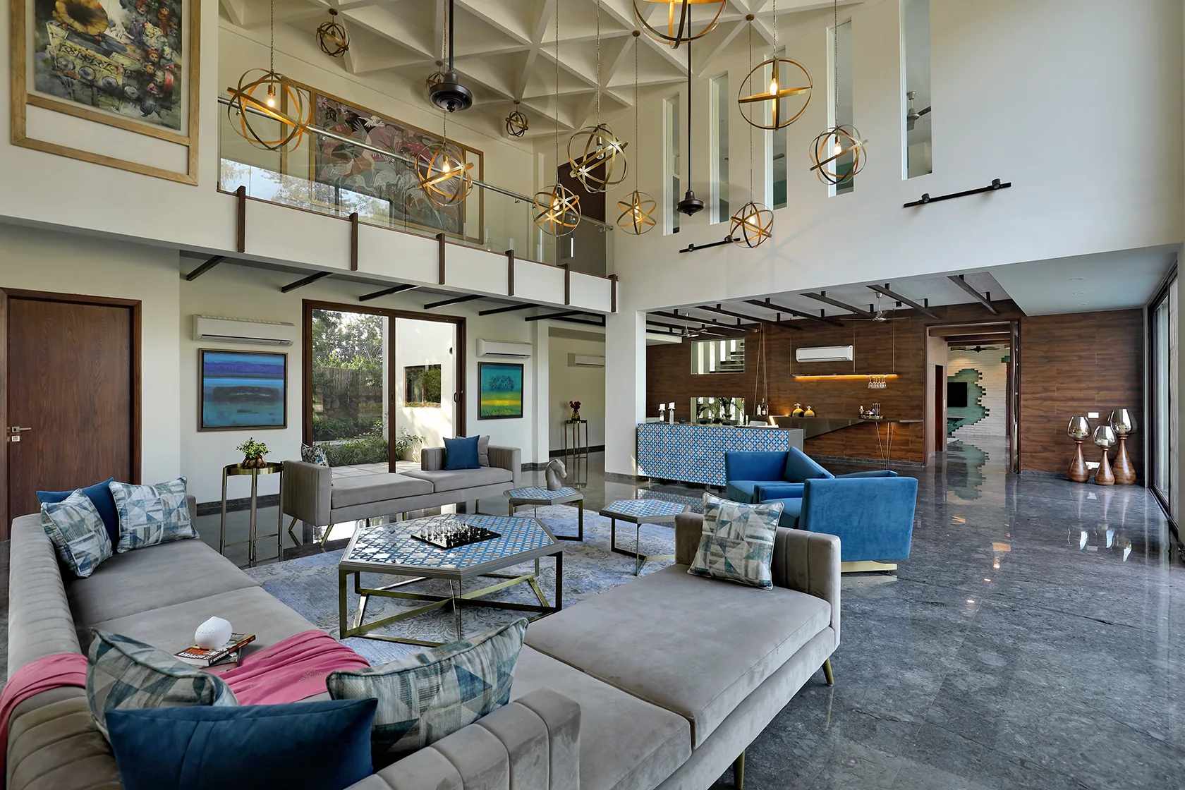 A large living room with grey sofas, blue accent chairs, a geometric coffered ceiling, hanging globe pendant lights, and polished stone flooring.