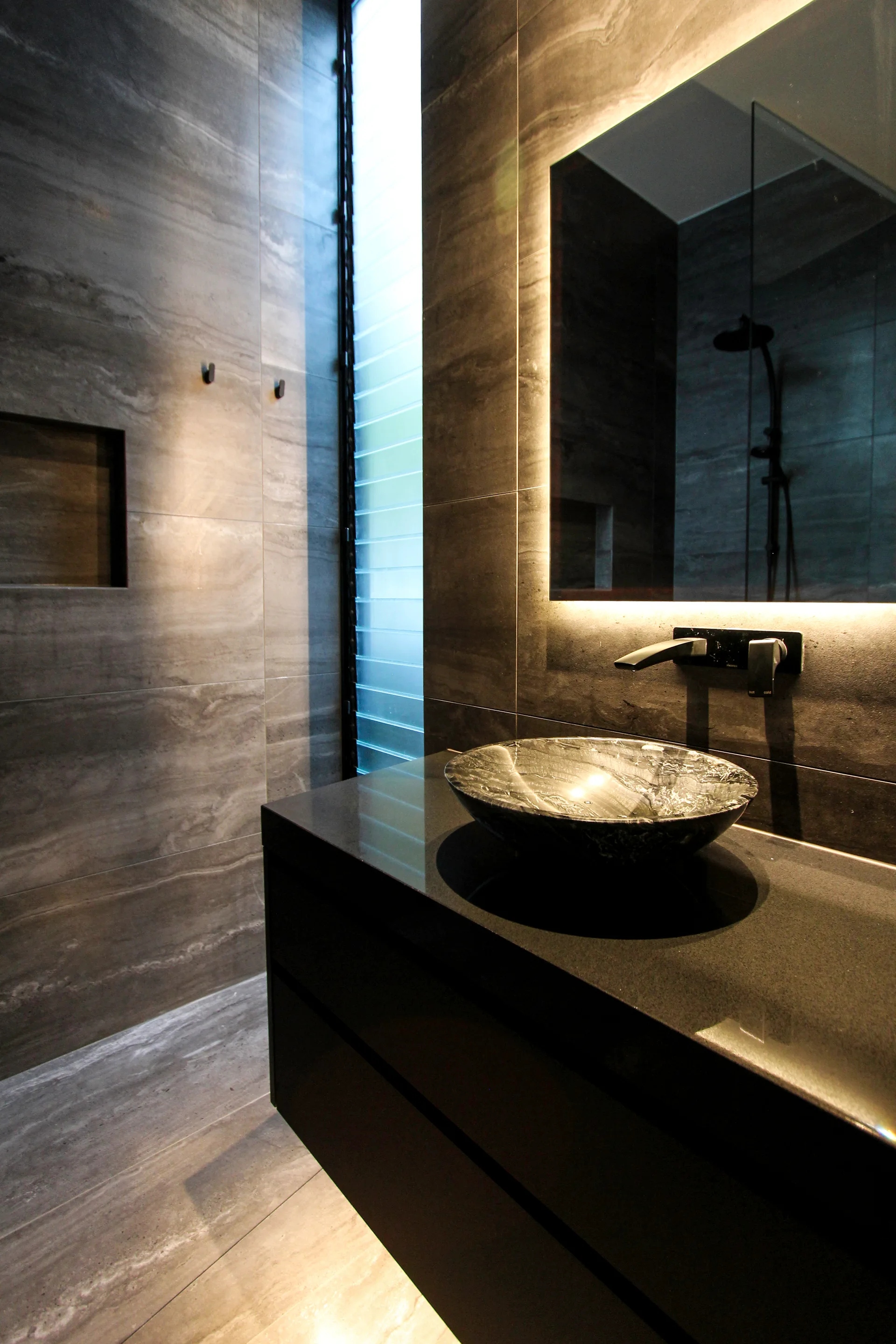 A modern grey stone bathroom featuring a black wall-mounted vanity, a circular vessel sink, and a backlit rectangular mirror.