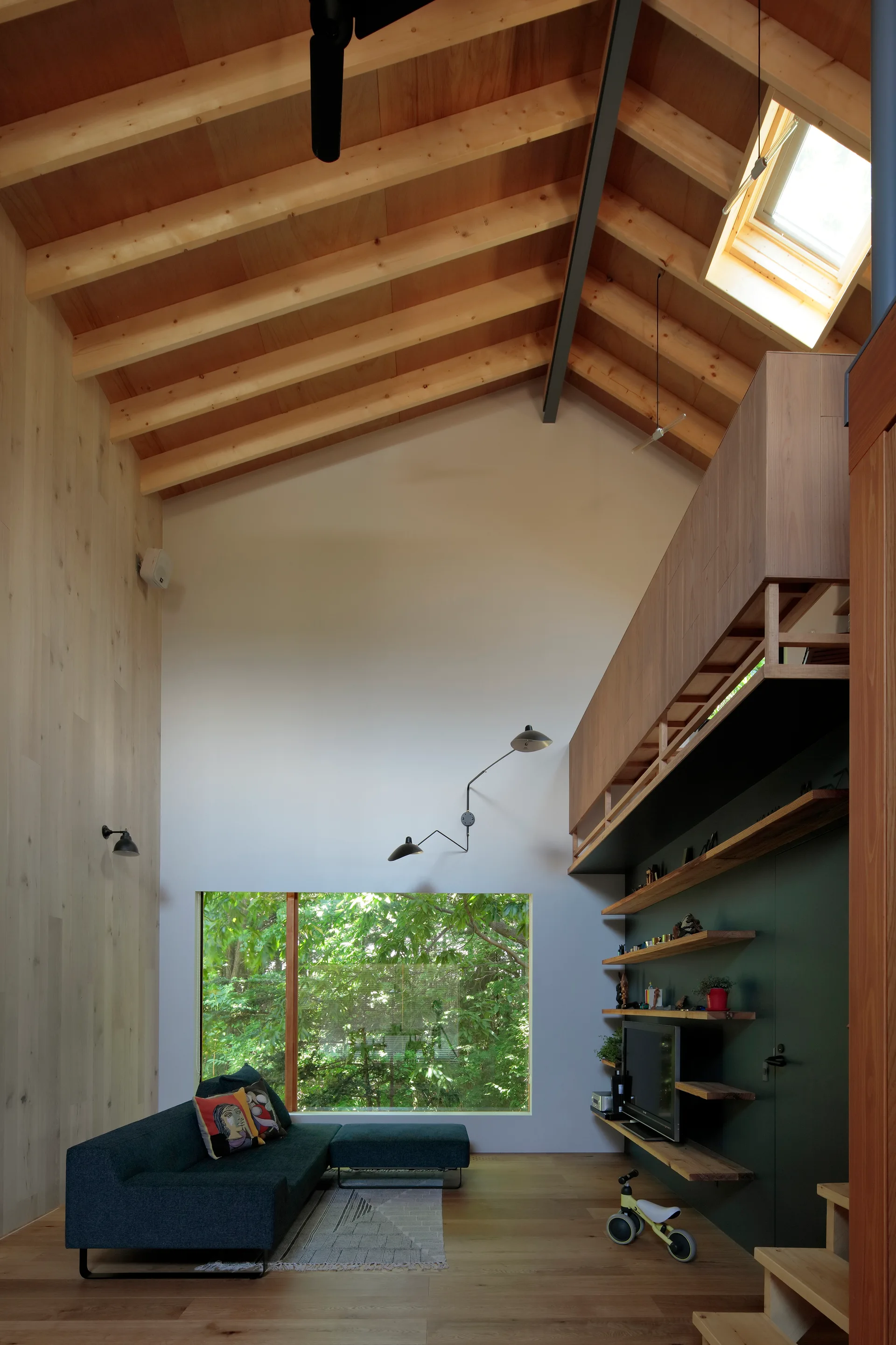 A double-height living room features a vaulted timber ceiling, a large landscape window looking out to green trees, and a dark blue sofa.