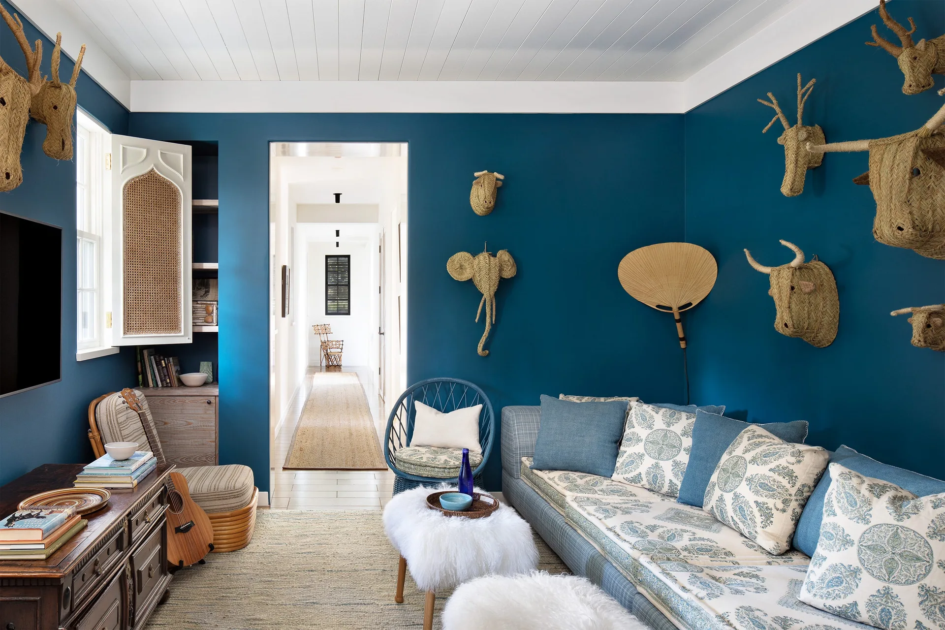 A vibrant blue living room features woven animal heads on the walls, a patterned daybed sofa, and white faux fur ottomans under a wood paneled ceiling.