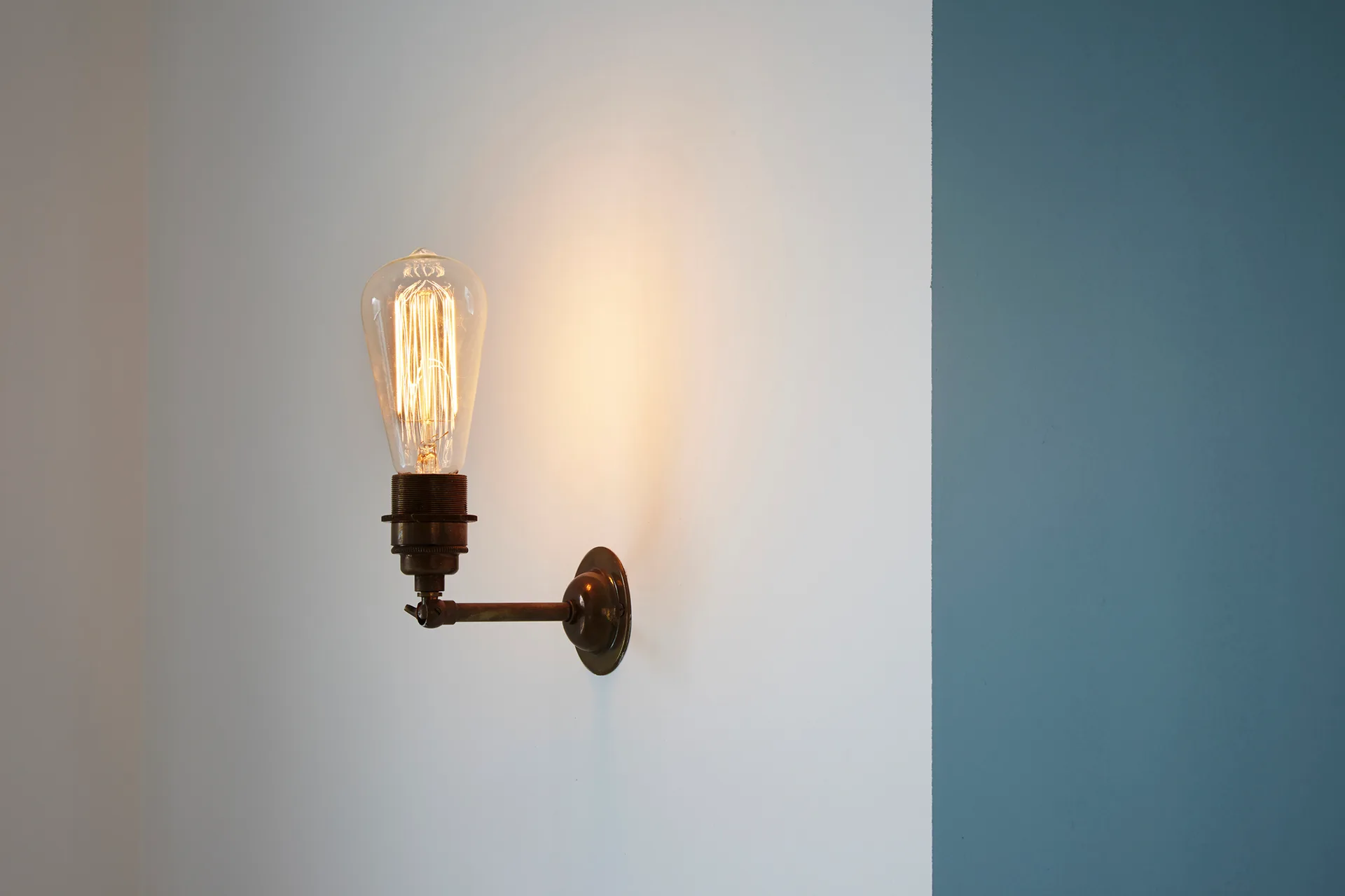A brass wall-mounted sconce with a glowing Edison-style filament bulb is affixed to a white painted wall beside a blue corner.