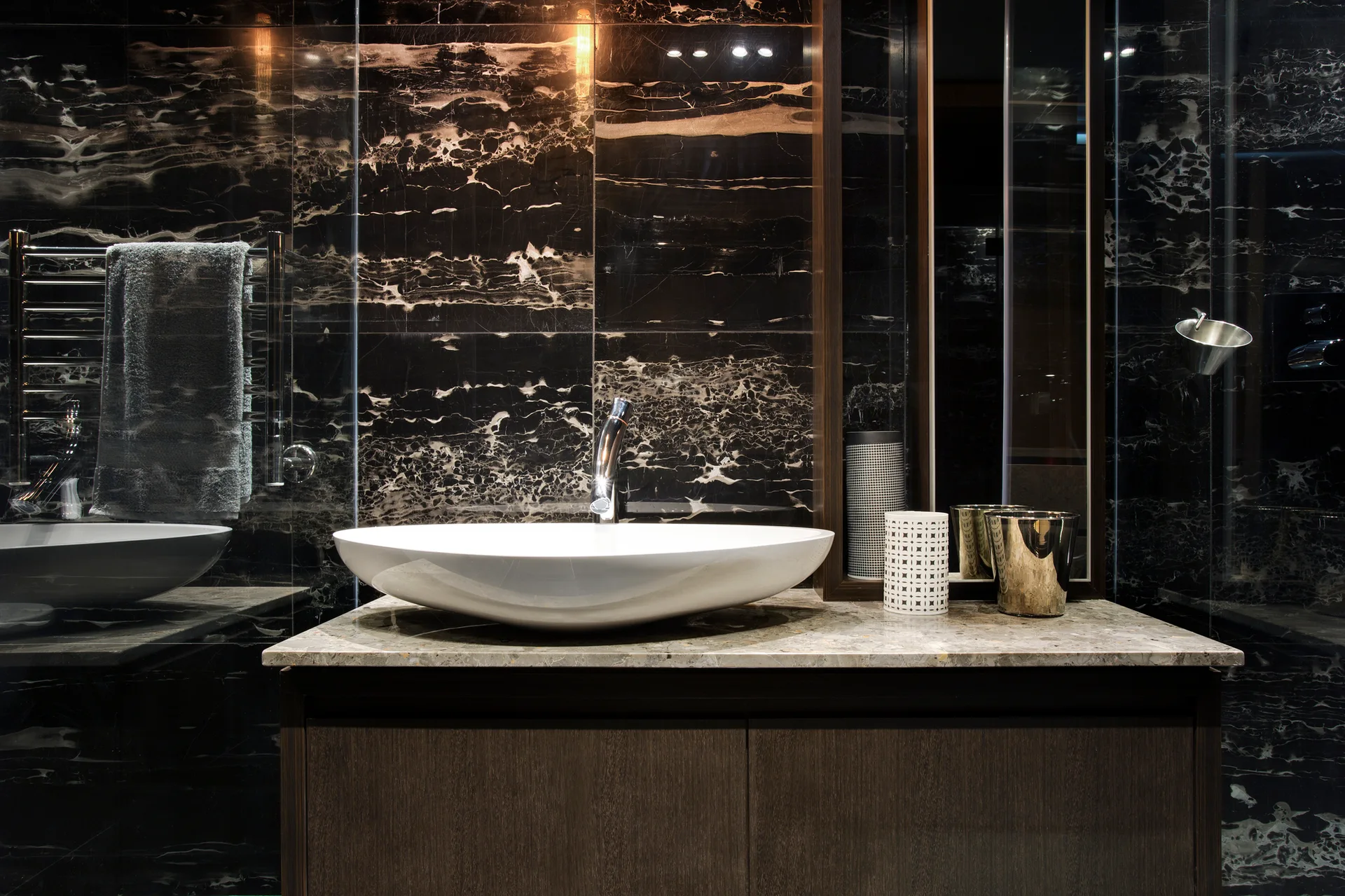 A modern bathroom vanity features a white oval vessel sink on a stone countertop against a polished black veined marble tiled wall.