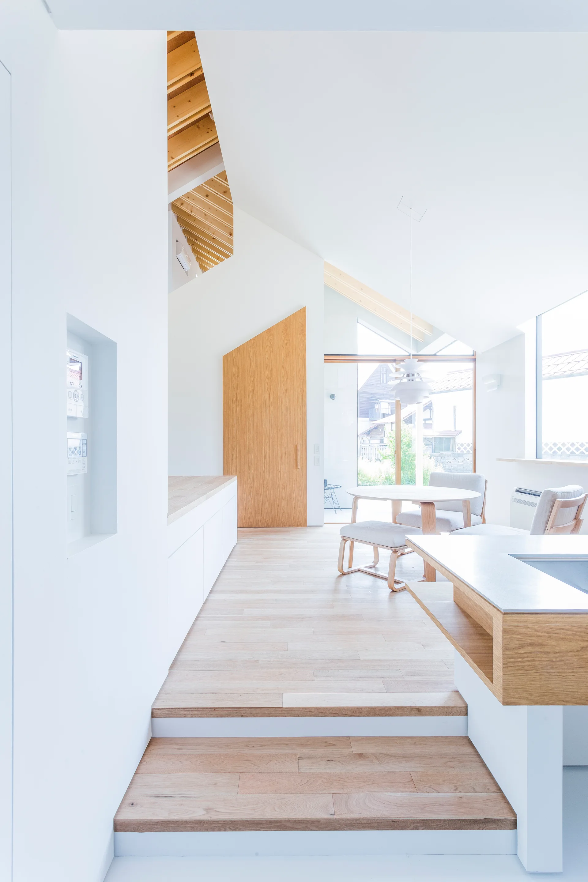 A minimalist dining and kitchen area featuring a stepped light wood floor, white walls, and a vaulted ceiling with exposed rafters.