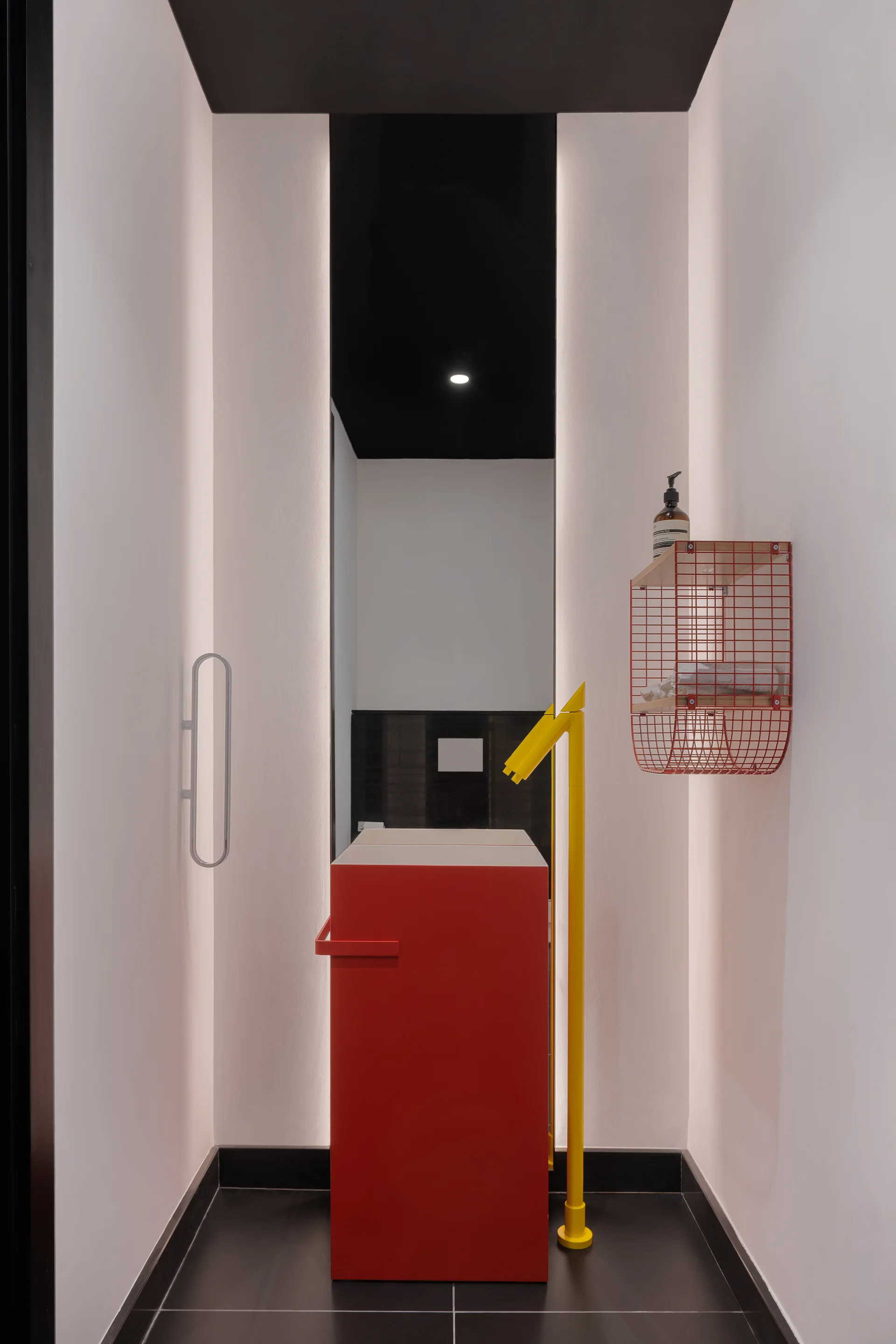 A narrow modernist powder room features a bright red block vanity, a yellow freestanding faucet, vertical cove lighting, and a black ceiling.