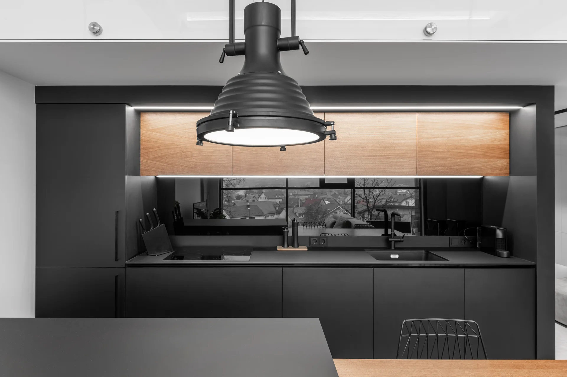 A modern kitchen with matte black cabinetry, warm wood upper cabinets, a mirrored backsplash, and a black industrial pendant light.