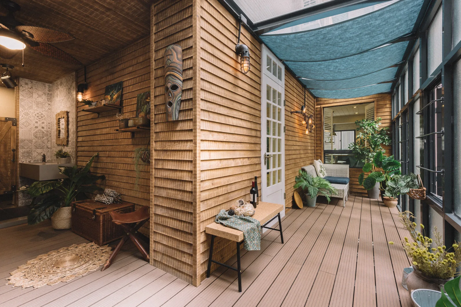 A covered wooden terrace features horizontal wood siding, a blue fabric overhead canopy, a white glass door, and potted green plants.