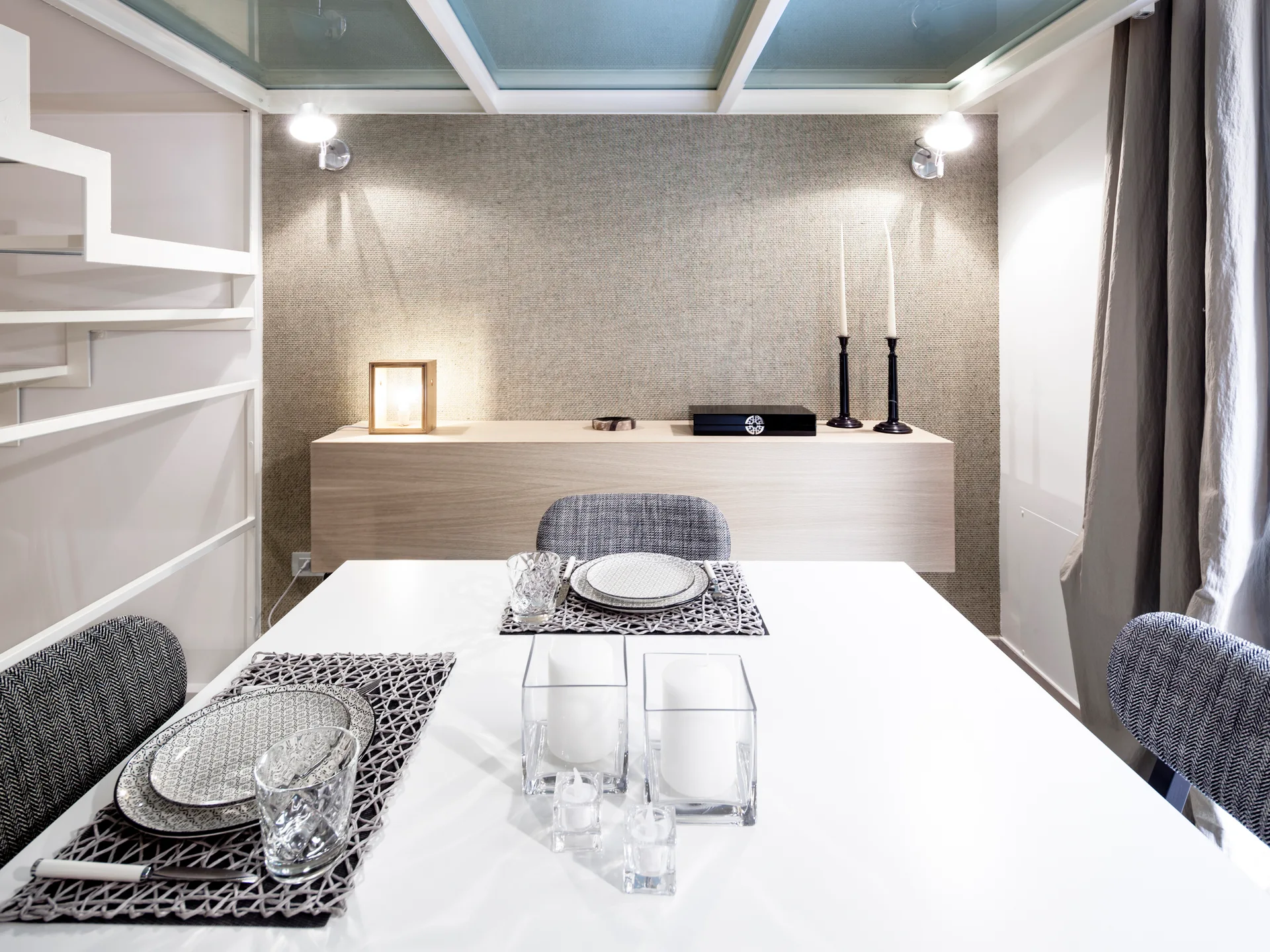 A white dining table is set with grey placemats in front of a floating wood sideboard and a textured beige accent wall.