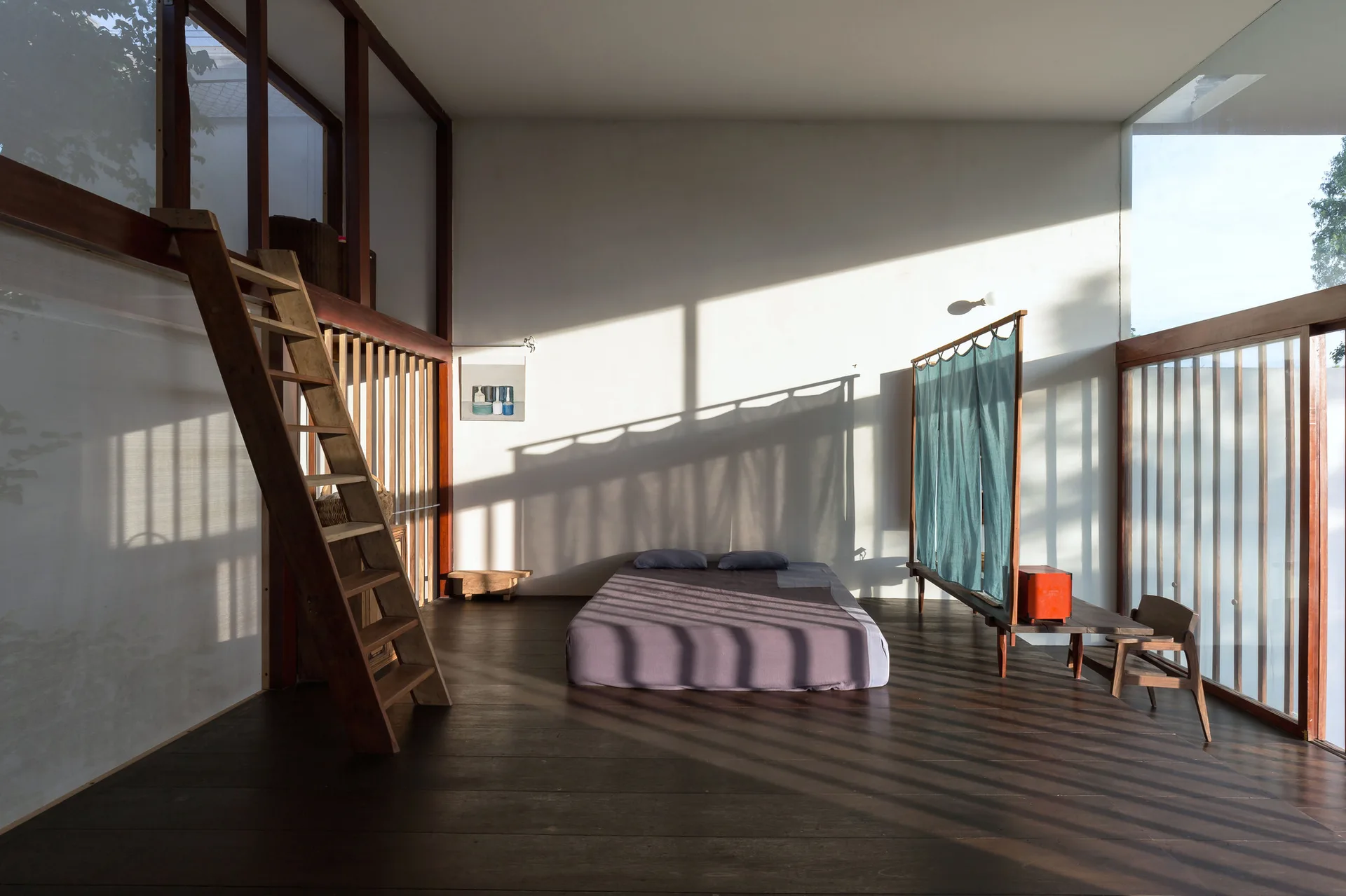 A minimalist bedroom featuring a low-profile bed on a dark wood floor, a wooden ladder, and a teal fabric screen under natural sunlight.