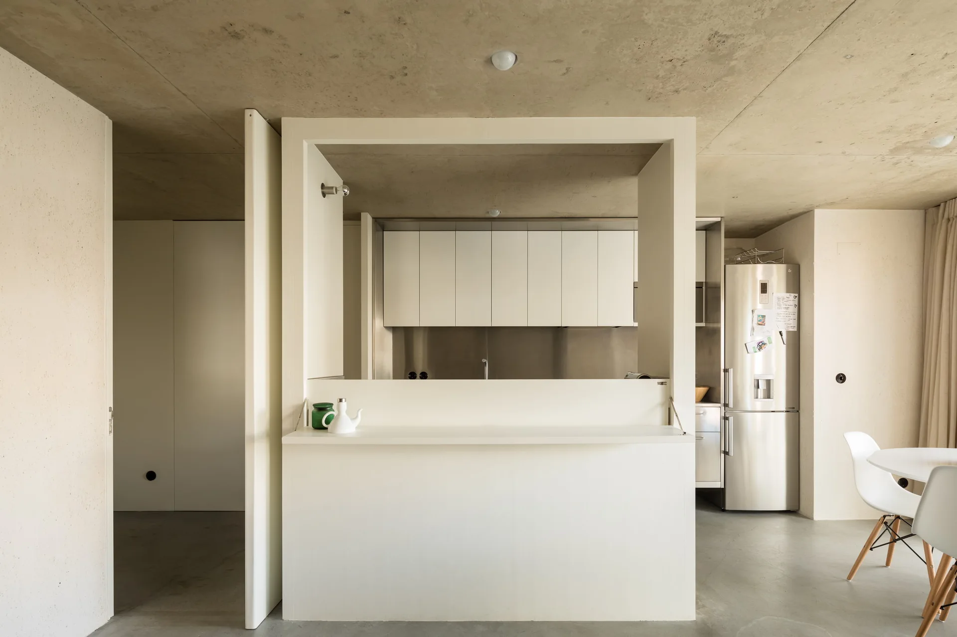 A minimalist kitchen features a white partition wall with a large pass-through opening under an exposed concrete ceiling with a dining table nearby.