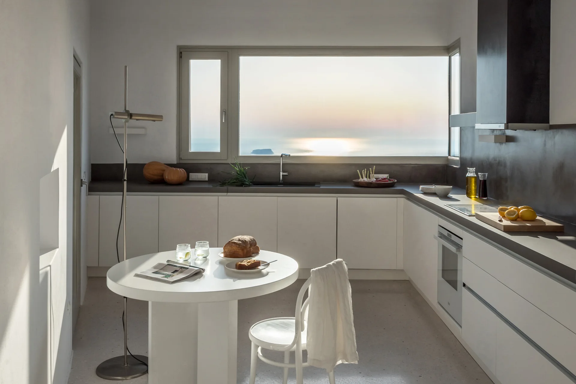 A minimalist white kitchen features a round pedestal table, gray countertops, and a wide horizontal window framing a coastal sunset over the ocean.