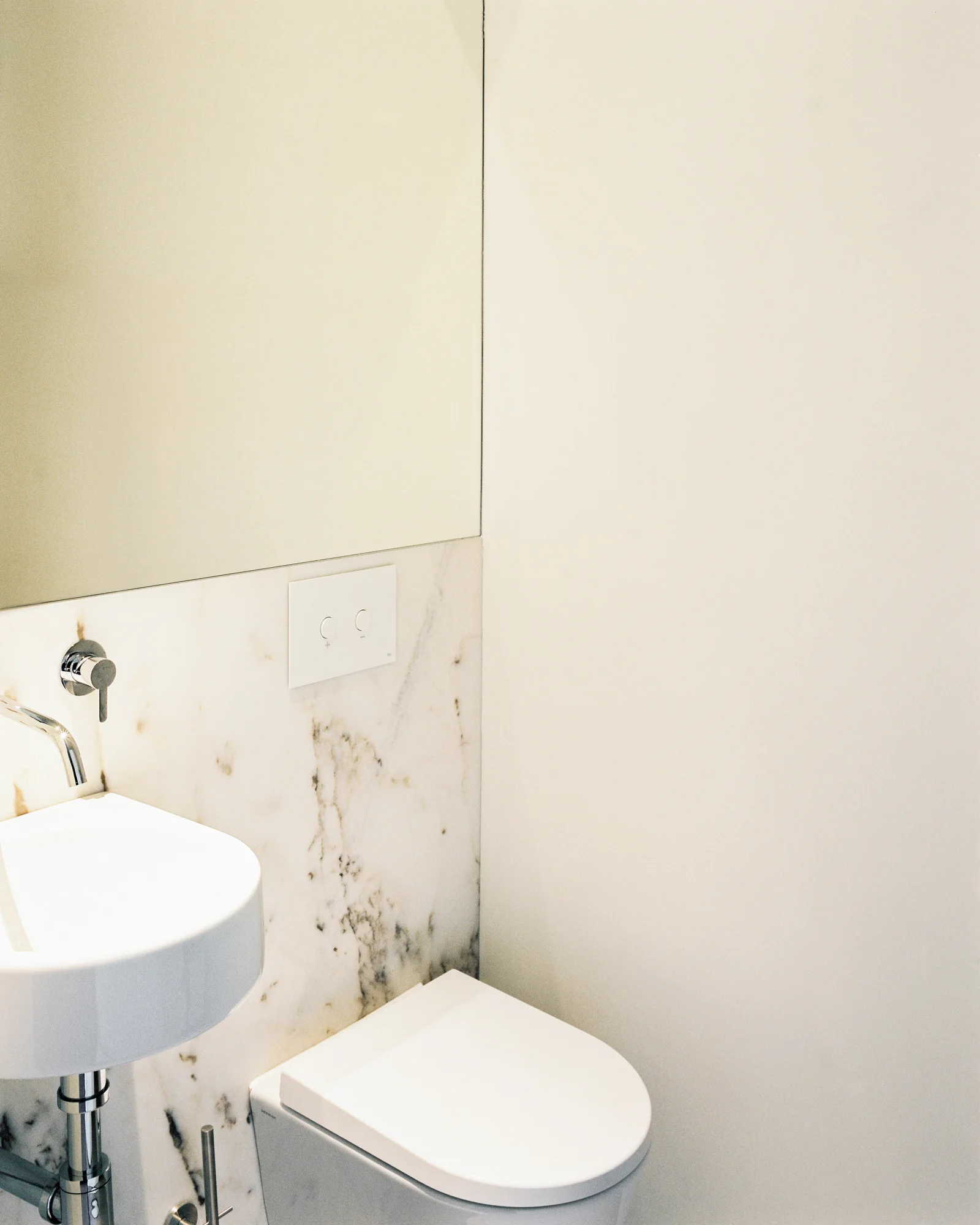 A minimalist powder room features a white wall-hung toilet, a small wall-mounted sink, and a veined marble backsplash beneath a large mirror.