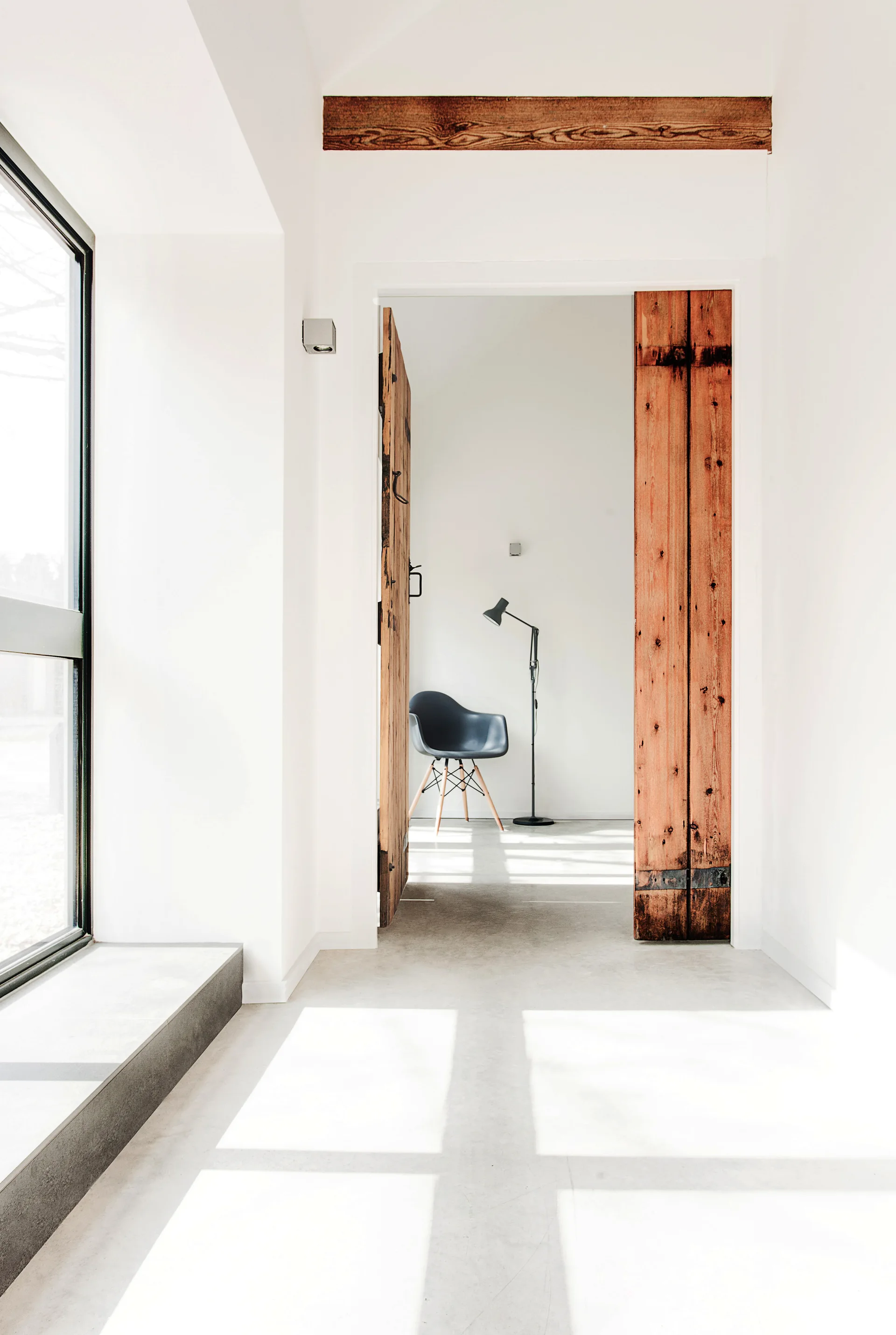 A bright minimalist hallway with white walls and concrete floors leads to an open reclaimed wood door, revealing a chair and lamp in the room beyond.