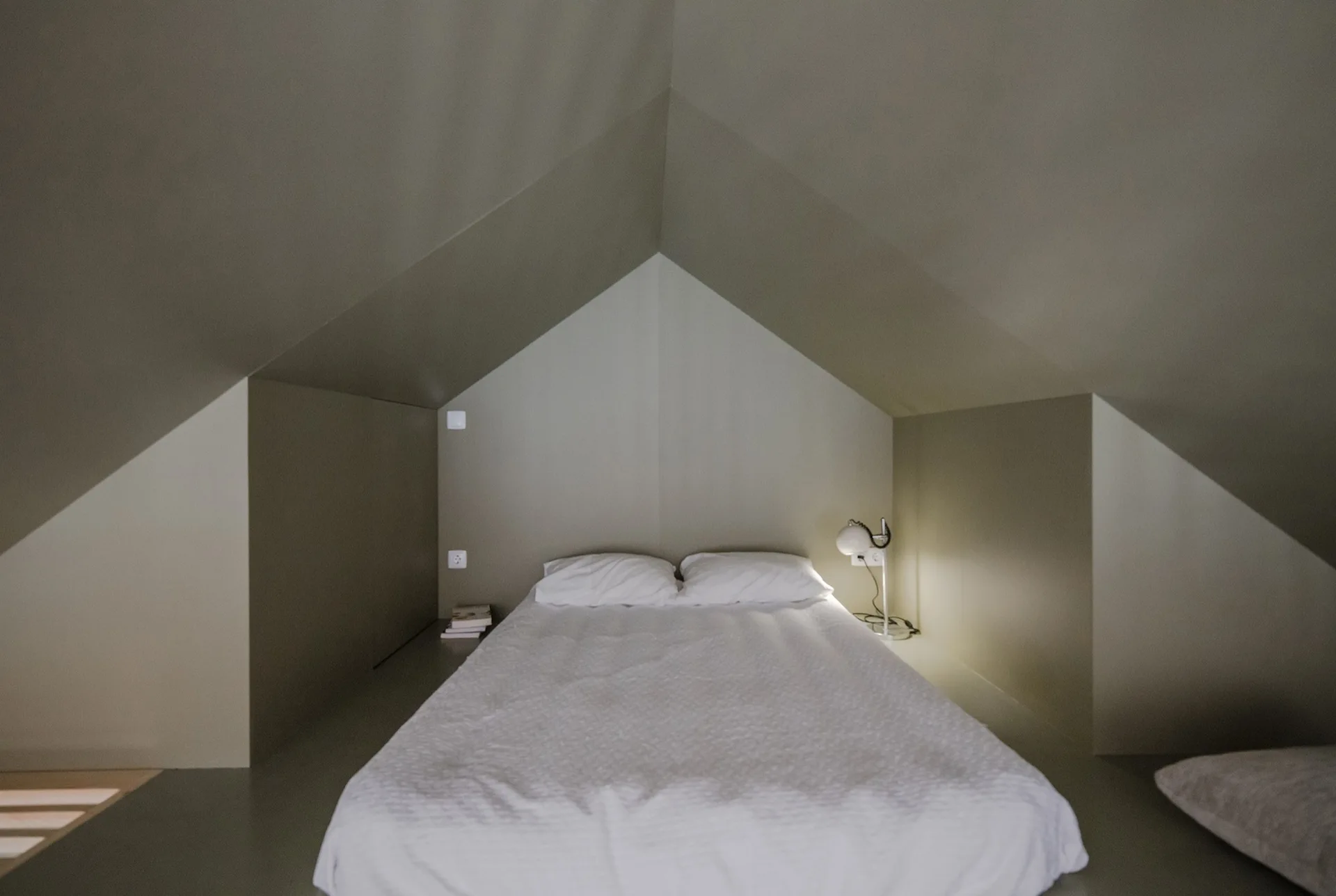 A bed with white bedding is nestled into a tight attic niche under a steeply pitched grey ceiling next to a small lit lamp.