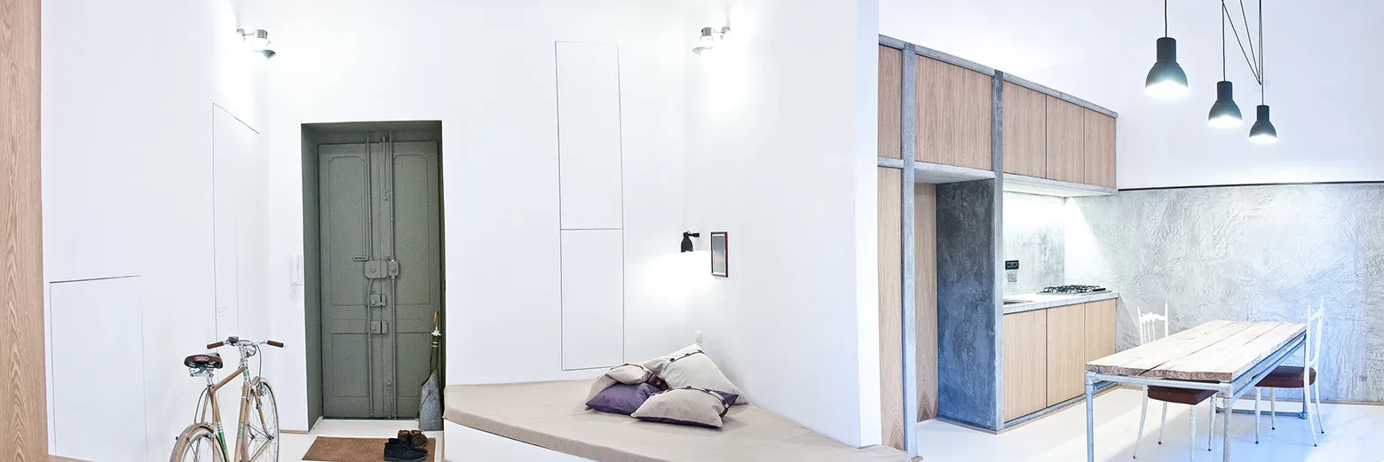 A wide-angle view of a minimalist open-plan apartment featuring a grey entry door, a built-in daybed bench, and a light wood kitchen.